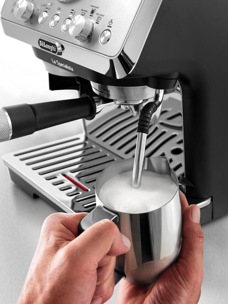Eine Espressomaschine schäumt Milch in einen Metallkrug. Hände halten den Krug.