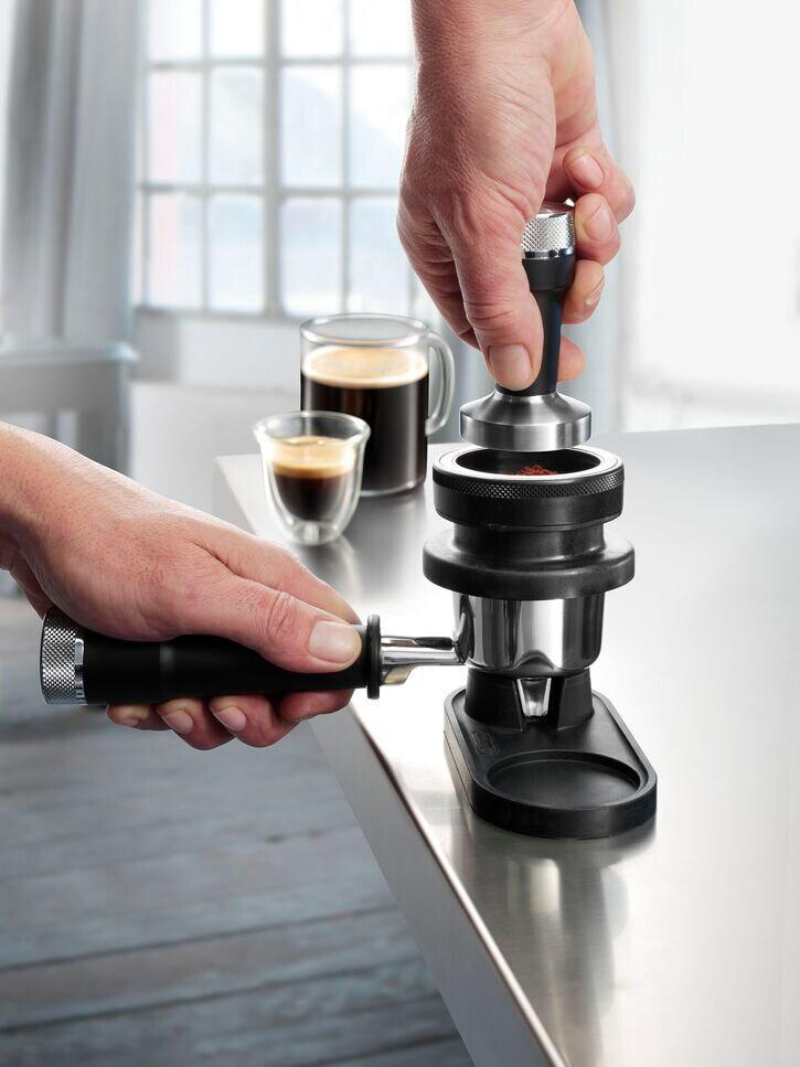 Hände, die Kaffeepulver mit einem schwarzen Tamper in einen Siebträger stopfen. Espressotassen im Hintergrund.