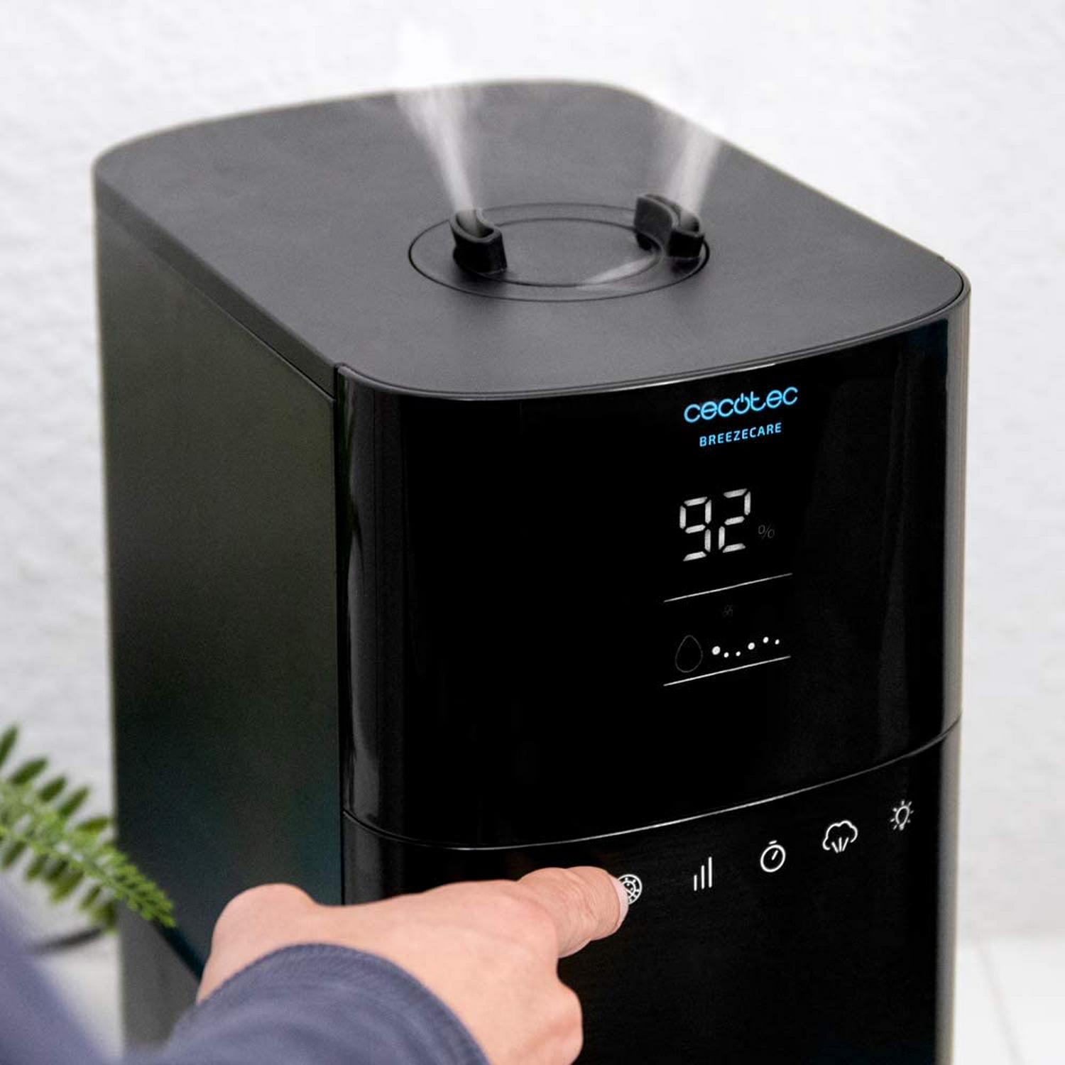 Una mano presiona un botón en un humidificador de aire negro. Una pantalla digital muestra 92%. La marca es Cecotec BREEZECARE.
