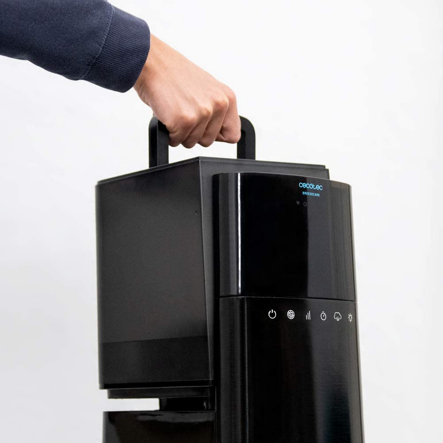 Una mano sostiene un purificador de aire negro con asa. La marca es Cecotec BreezeCare. Se ven algunos botones en el frente.