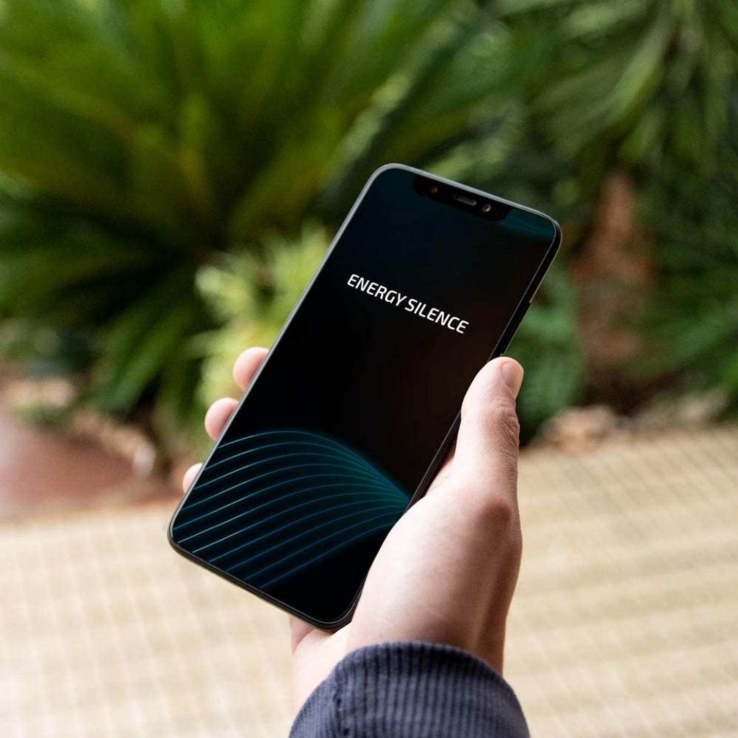 Una mano sostiene un smartphone con 'ENERGY SILENCE' en la pantalla. El follaje verde está borroso en el fondo. El teléfono tiene un diseño curvo.