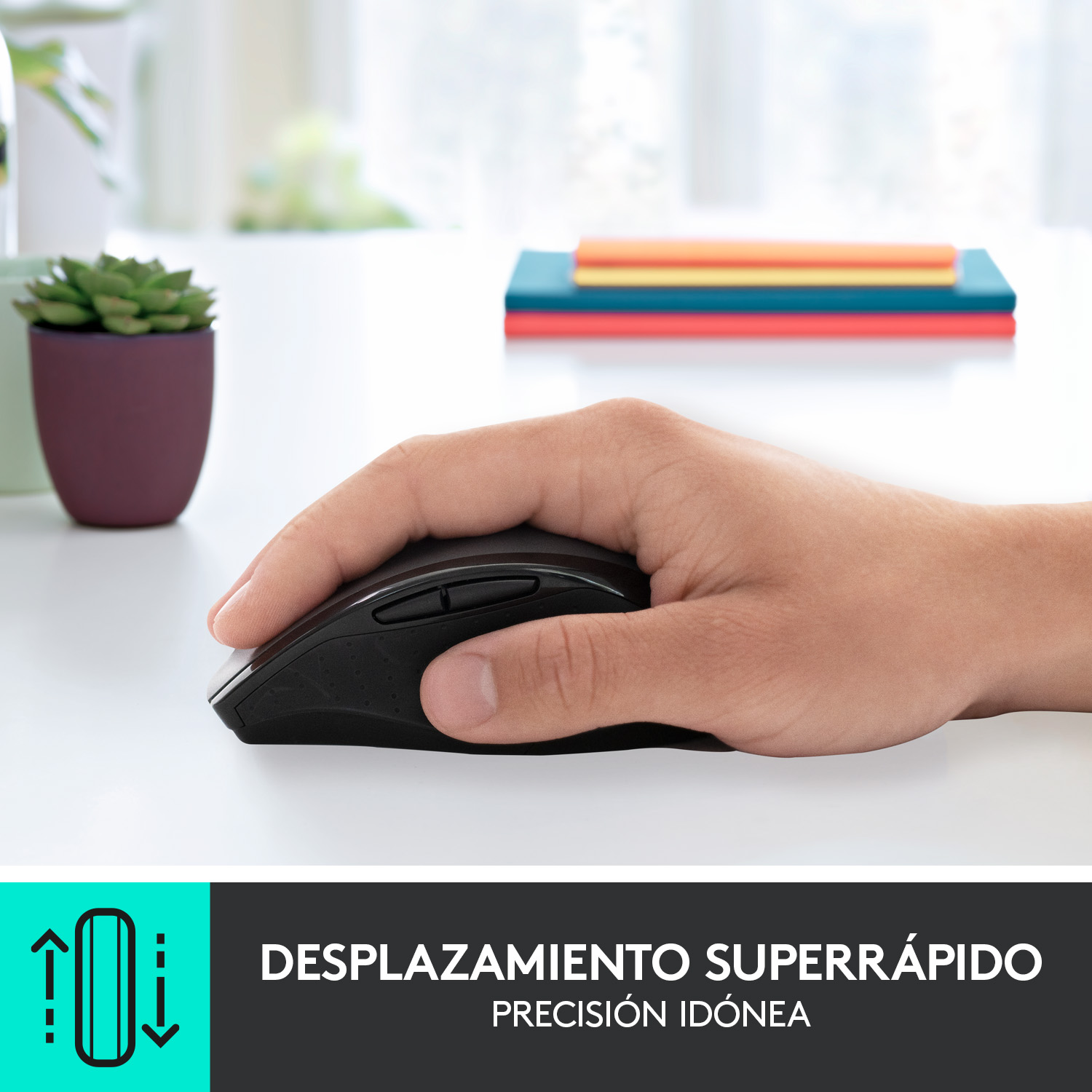 Una mano sosteniendo un ratón negro sobre una mesa blanca. Una planta pequeña y libros están en el fondo.