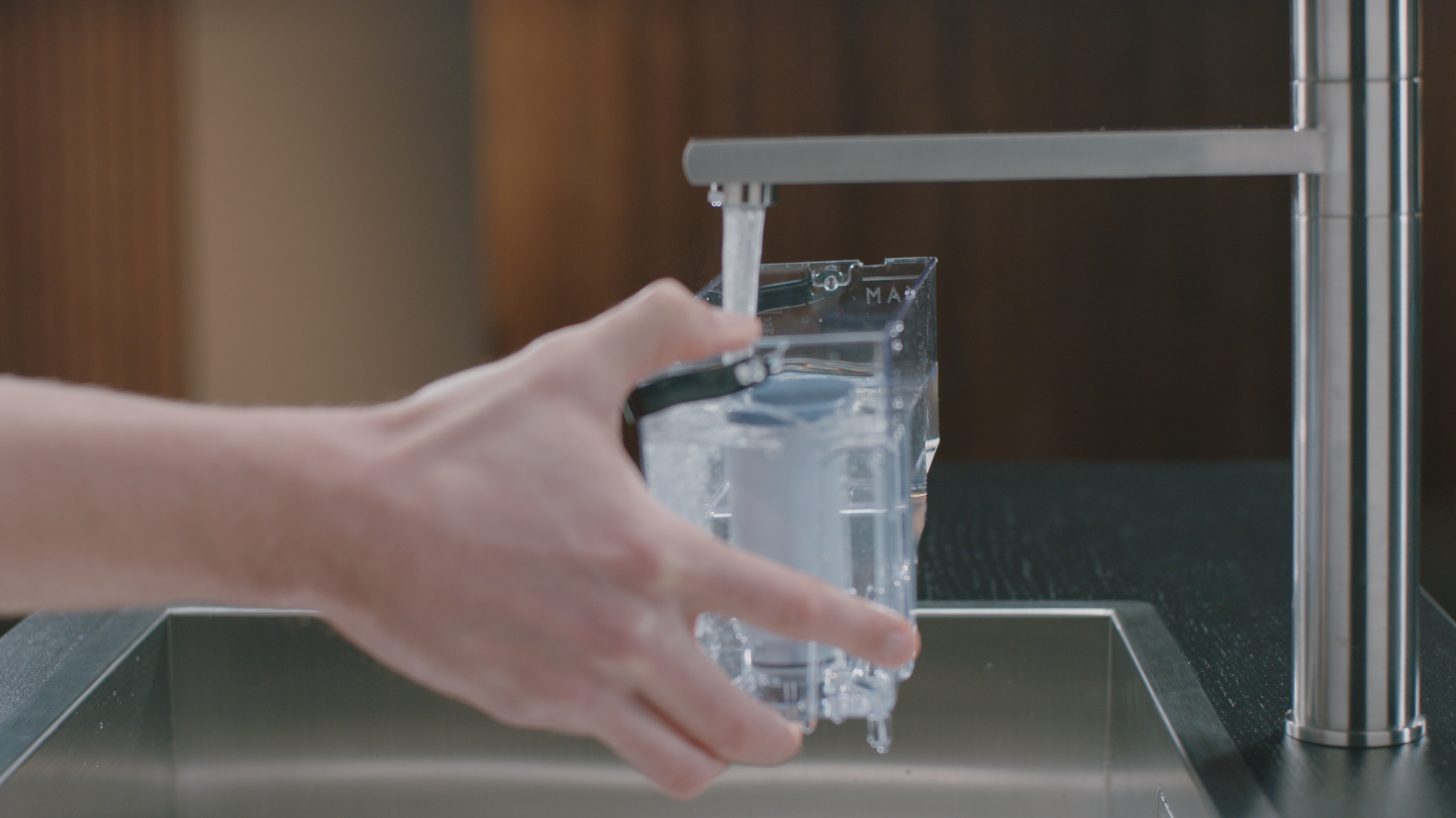 Hand filling a water filter container under a faucet in a kitchen setting, water pouring.