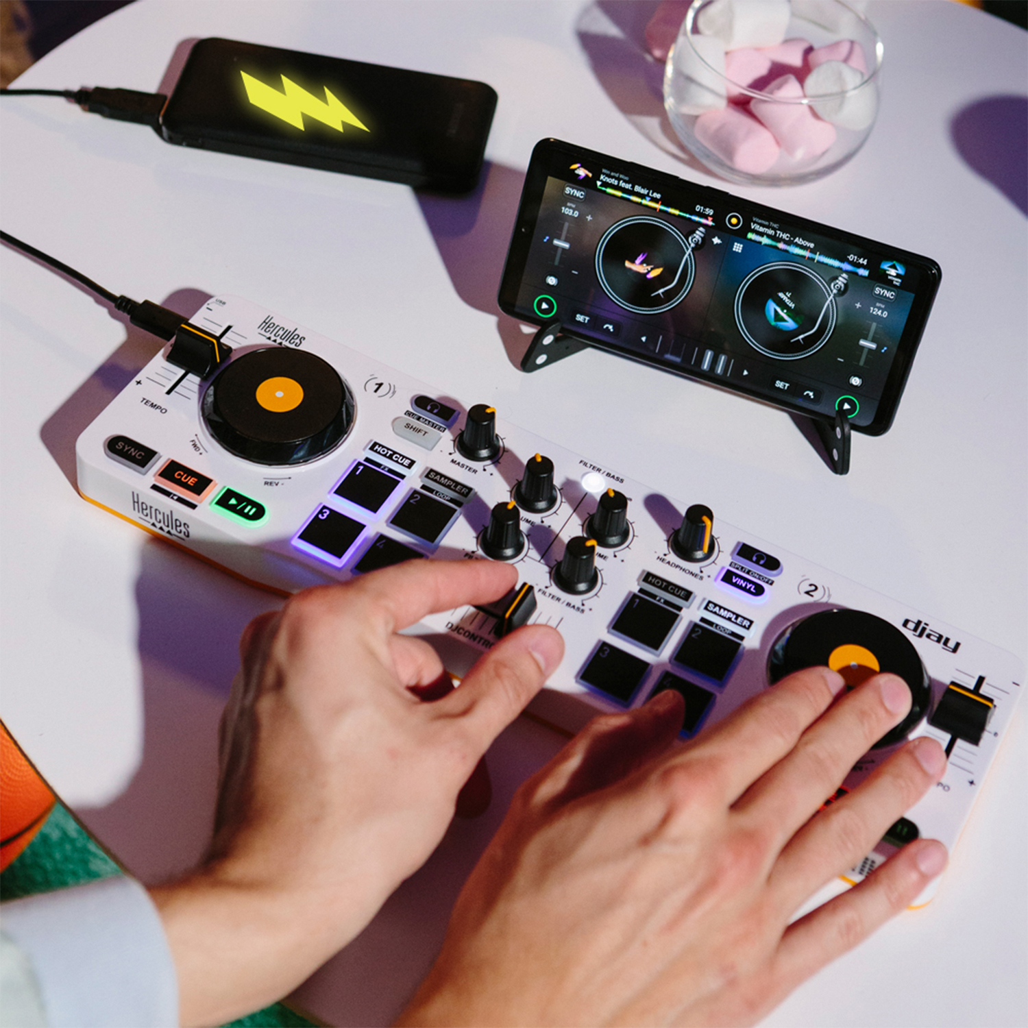 Controller DJ con smartphone che mostra un'interfaccia musicale. Le mani usano il controller. Un caricatore per telefono.