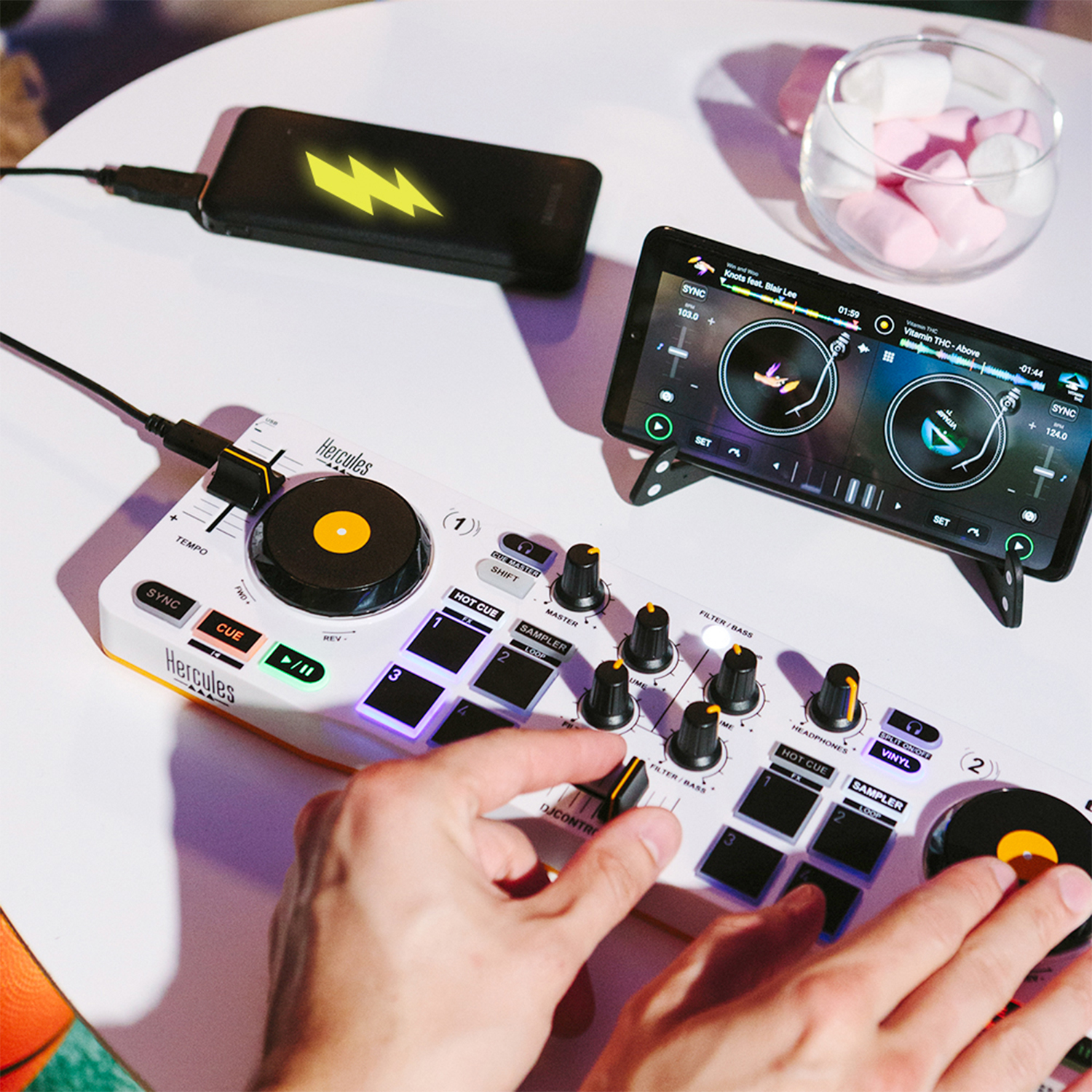 Un controller DJ con un telefono e un power bank su un tavolo bianco. Marshmallow nelle vicinanze.