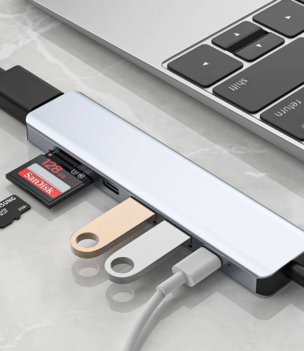 Çeşitli bağlı cihazlar ve arka planda bir dizüstü bilgisayar klavyesi bulunan gümüş USB hub.