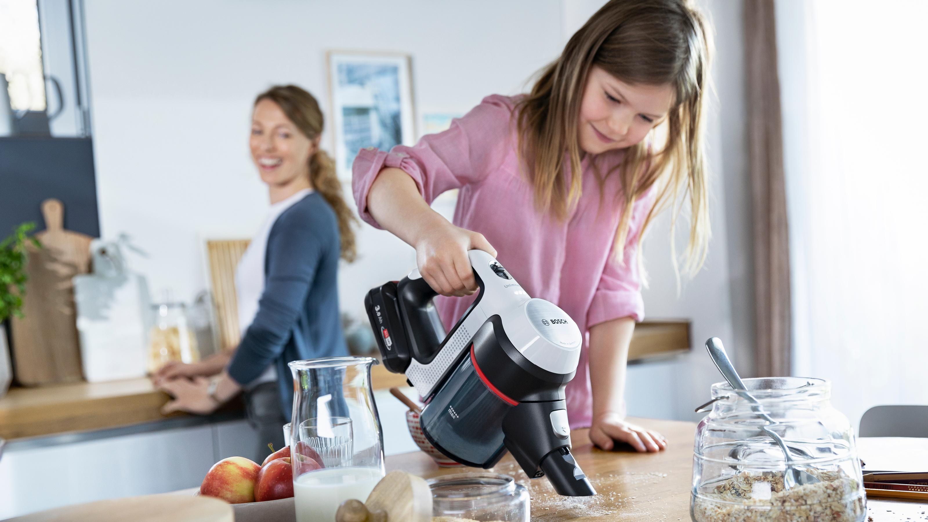 Ein Mädchen saugt einen Tisch. Eine Frau ist im Hintergrund. Küchenszene mit Lebensmitteln.