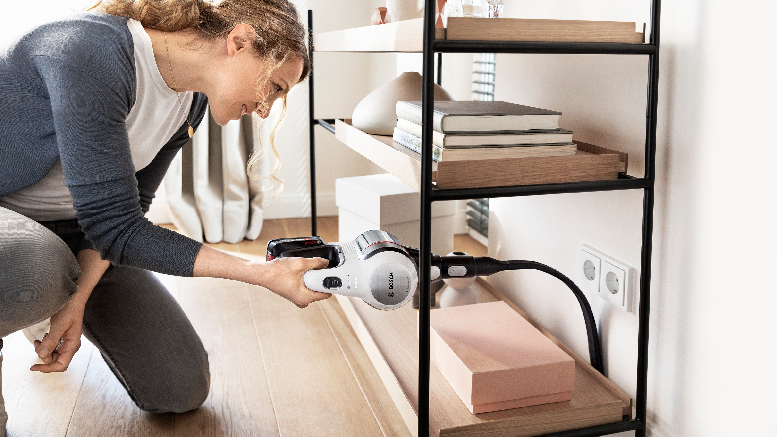 Frau reinigt Regal mit Staubsauger. Regal mit Büchern und Boxen. Holzfußboden davor.