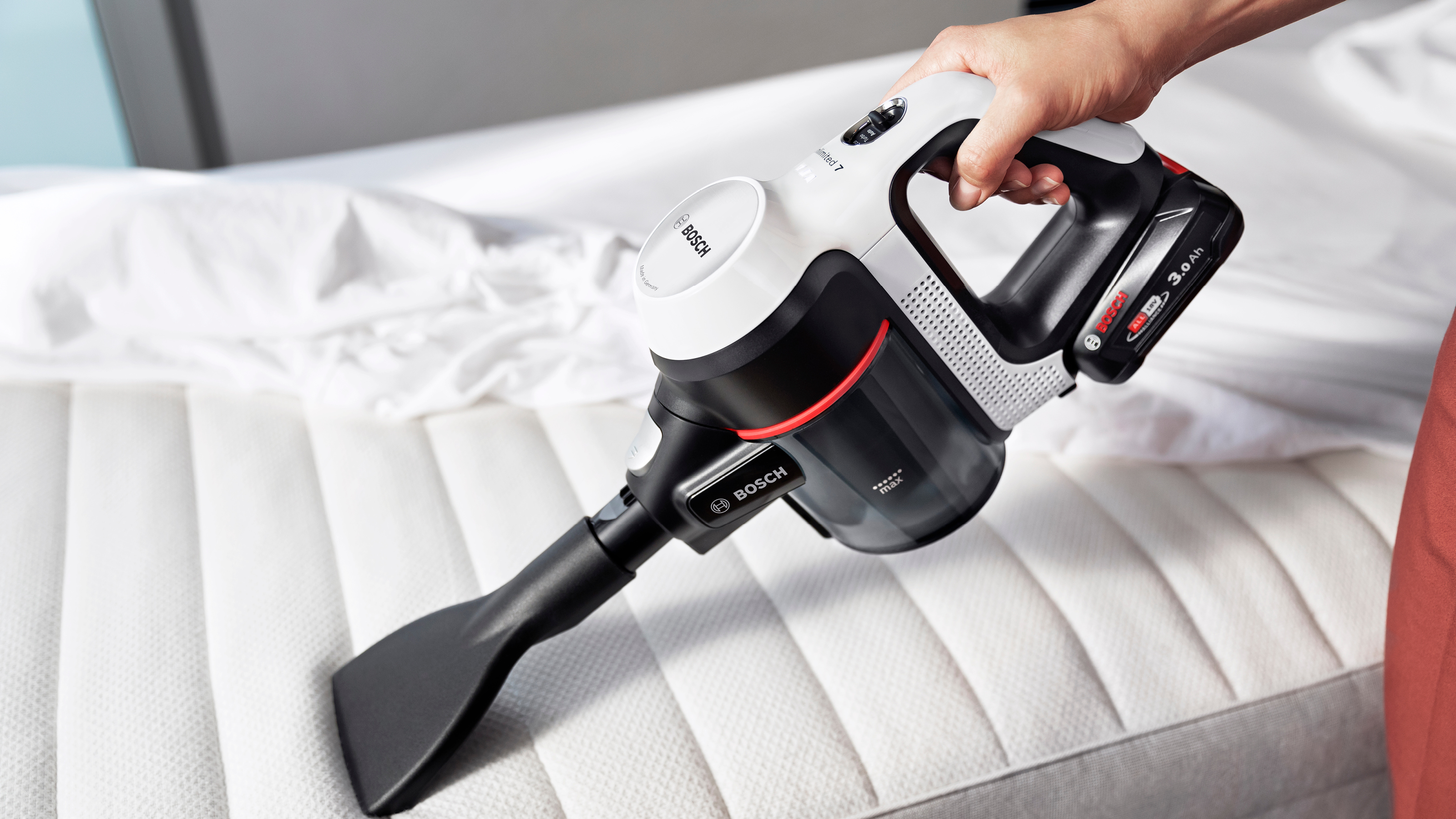 Person benutzt einen weißen und schwarzen Bosch Staubsauger auf einem weißen Bett.