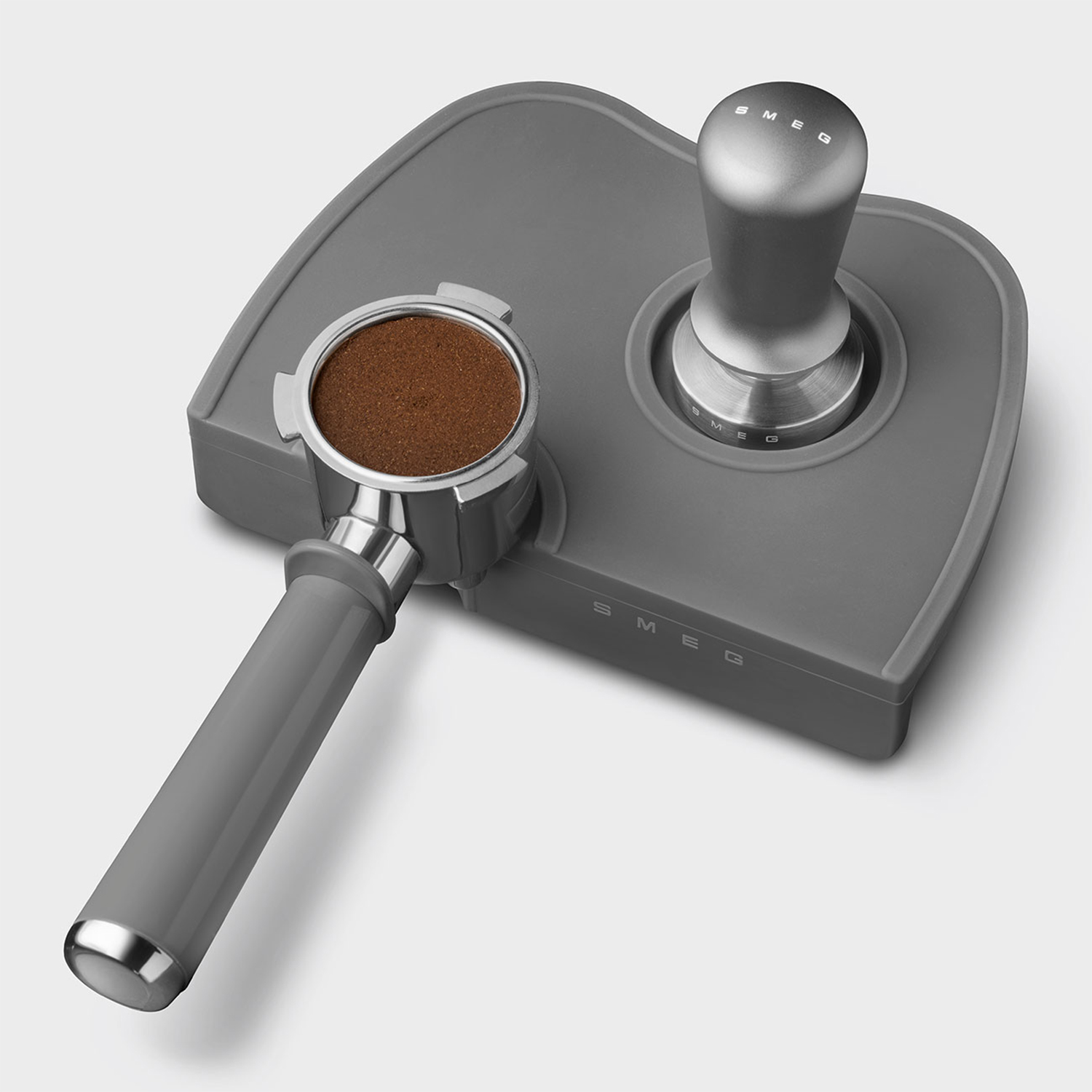 Pressino e portafiltro per caffè SMEG grigi su un tappetino grigio. Il portafiltro contiene caffè macinato.