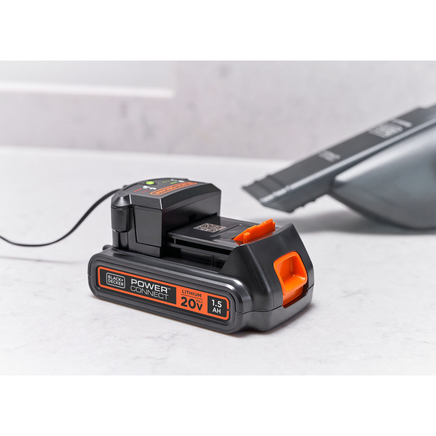 Caricabatterie Black+Decker nero e arancione su una superficie chiara con un aspirapolvere sullo sfondo.