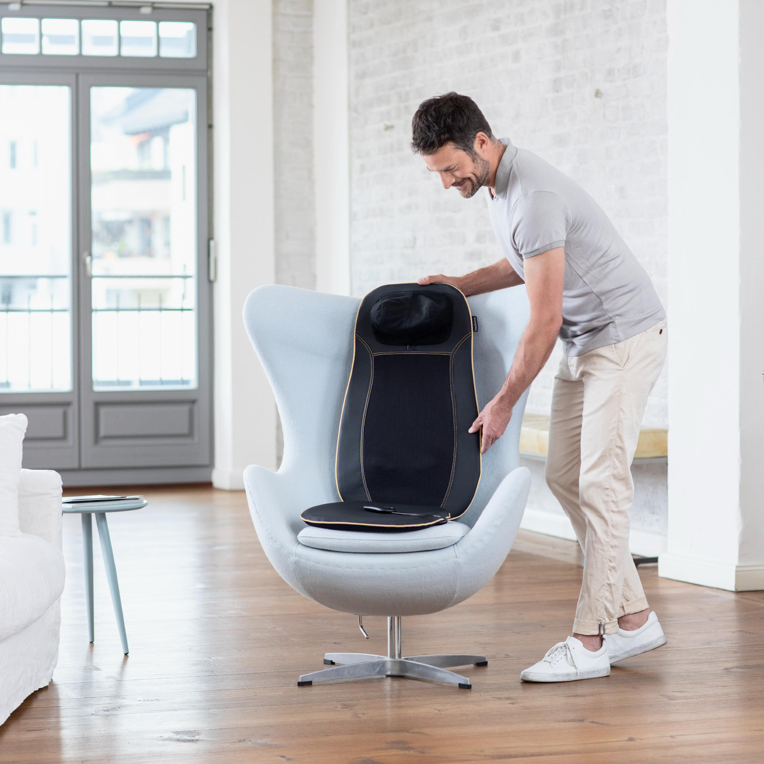 Ein Mann platziert einen schwarzen Massagesitz auf einem hellblauen Eiersessel in einem Wohnzimmer.