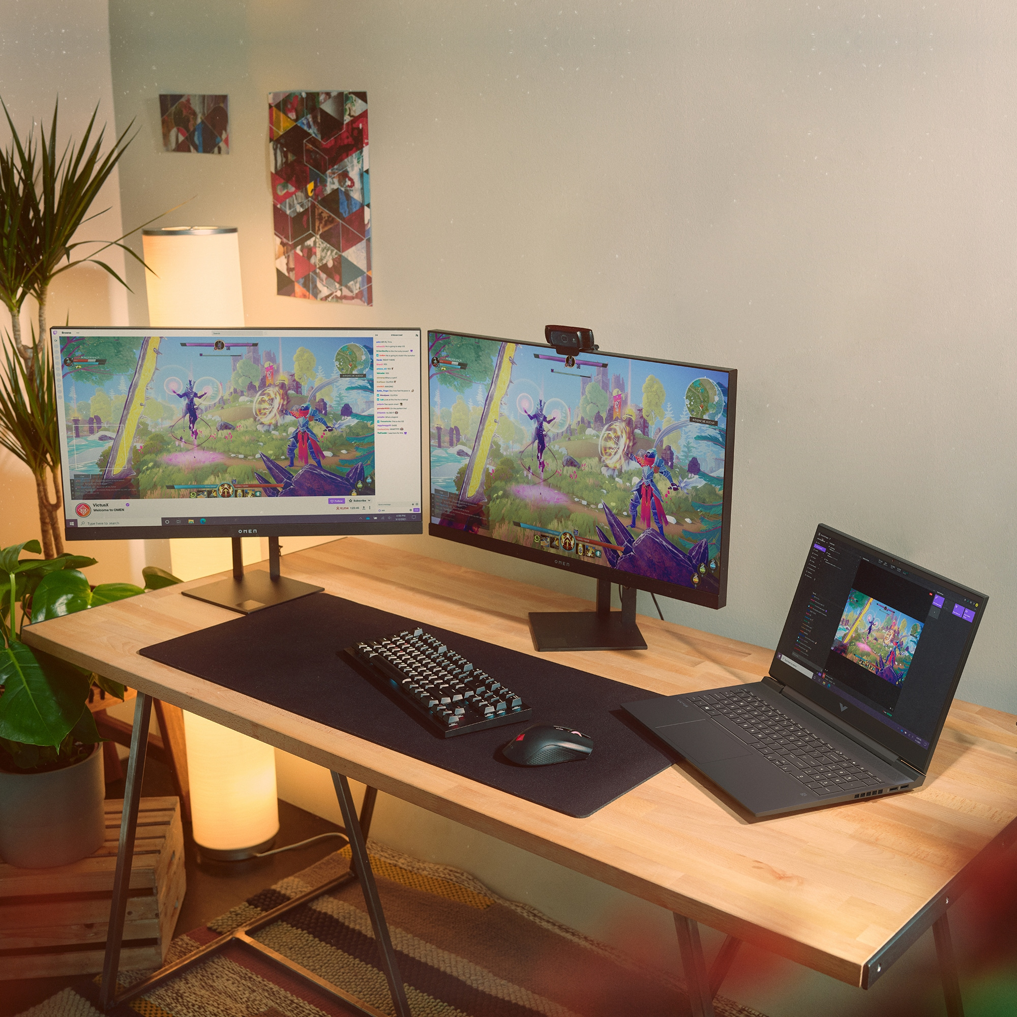 Dos monitores, teclado, portátil y plantas en un escritorio de madera. Una webcam está encima de un monitor.