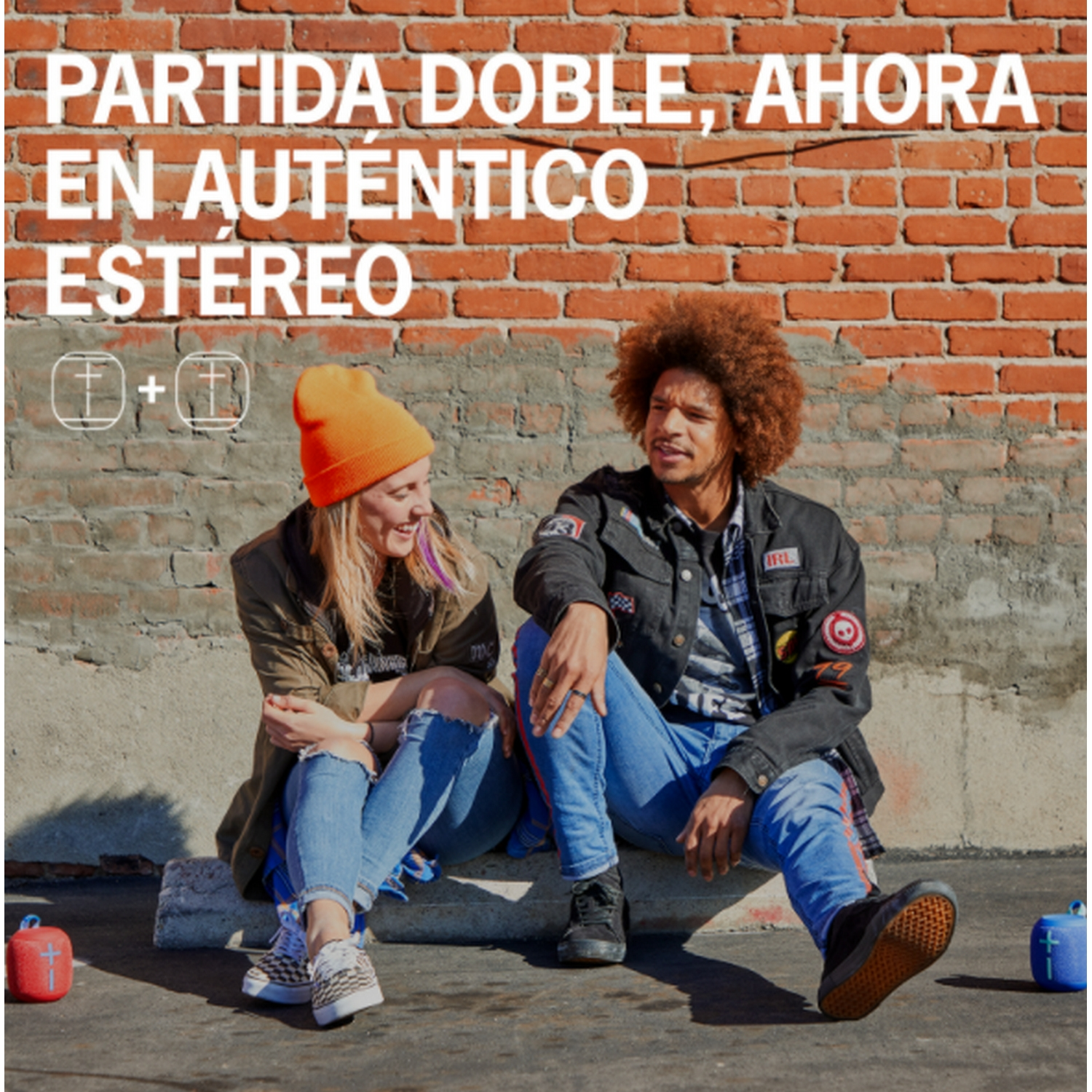 Dos personas sentadas, hablando, vestidas con ropa informal, junto a una pared de ladrillos.