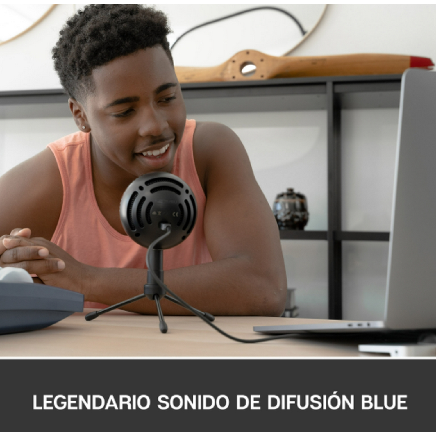 Un joven está sentado en un escritorio con un micrófono y una computadora portátil. Está sonriendo.