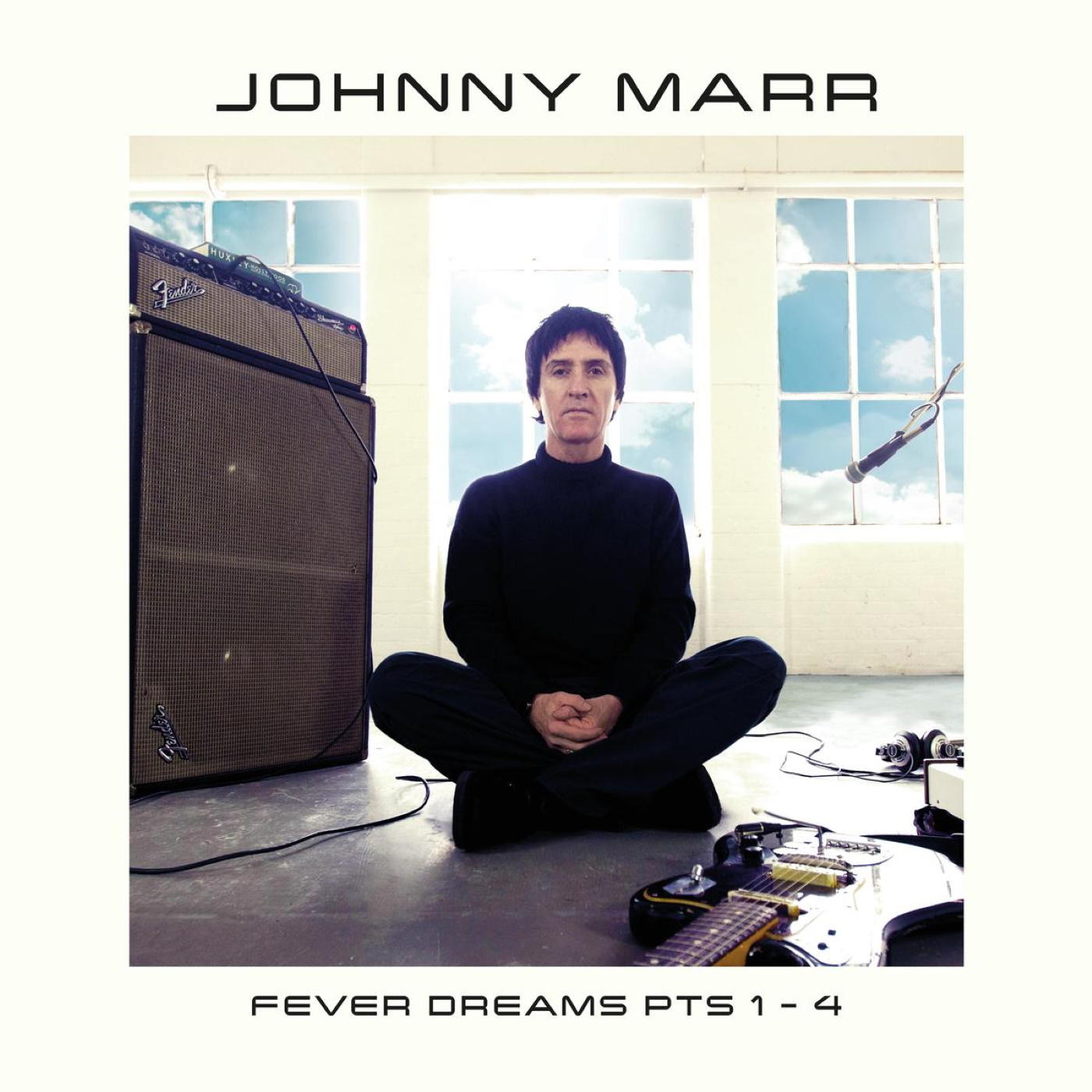 Johnny Marr assis avec une guitare, des vêtements noirs et un fond de fenêtres.