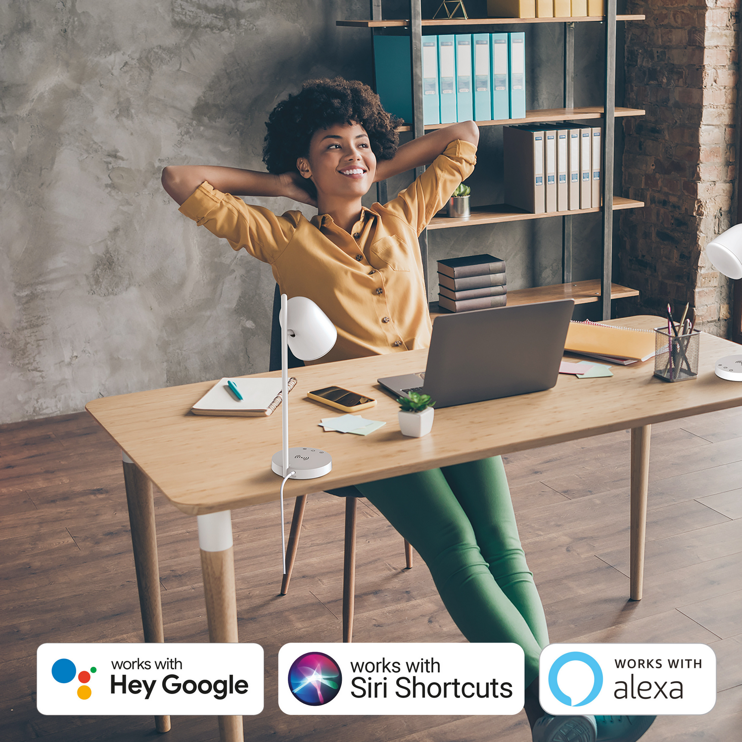 Mujer sentada en escritorio, sonriendo. Escritorio con portátil y lámpara. Logos: Hey Google, Siri, Alexa.