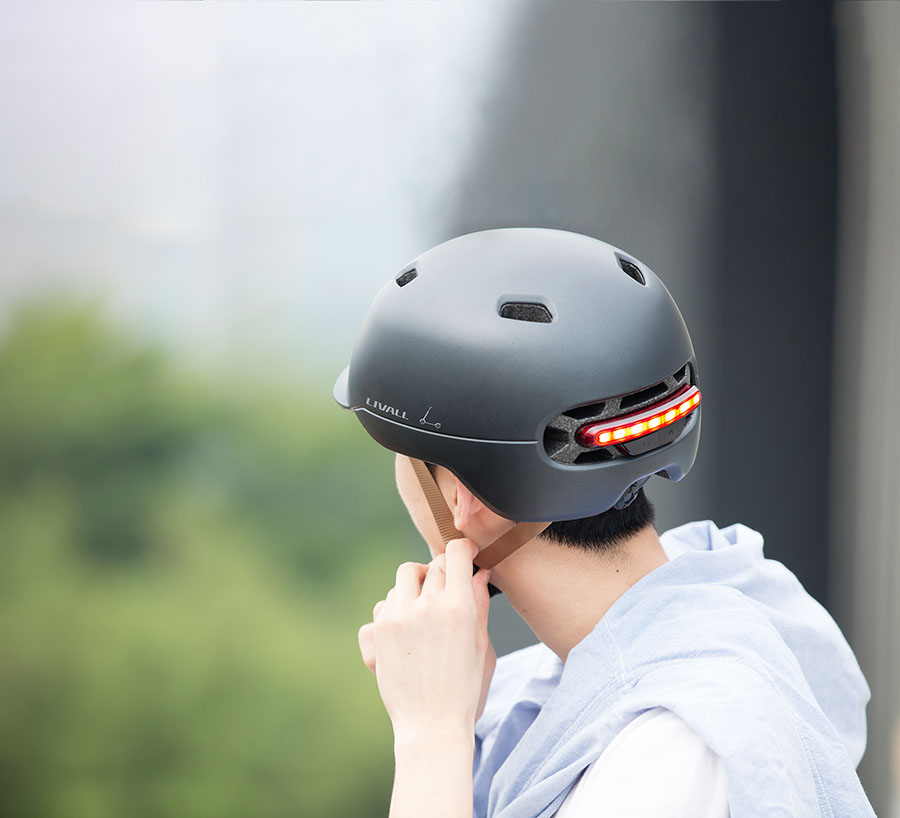 Una persona che indossa un casco da bicicletta grigio scuro con una luce rossa sul retro. Lo sfondo è sfocato.