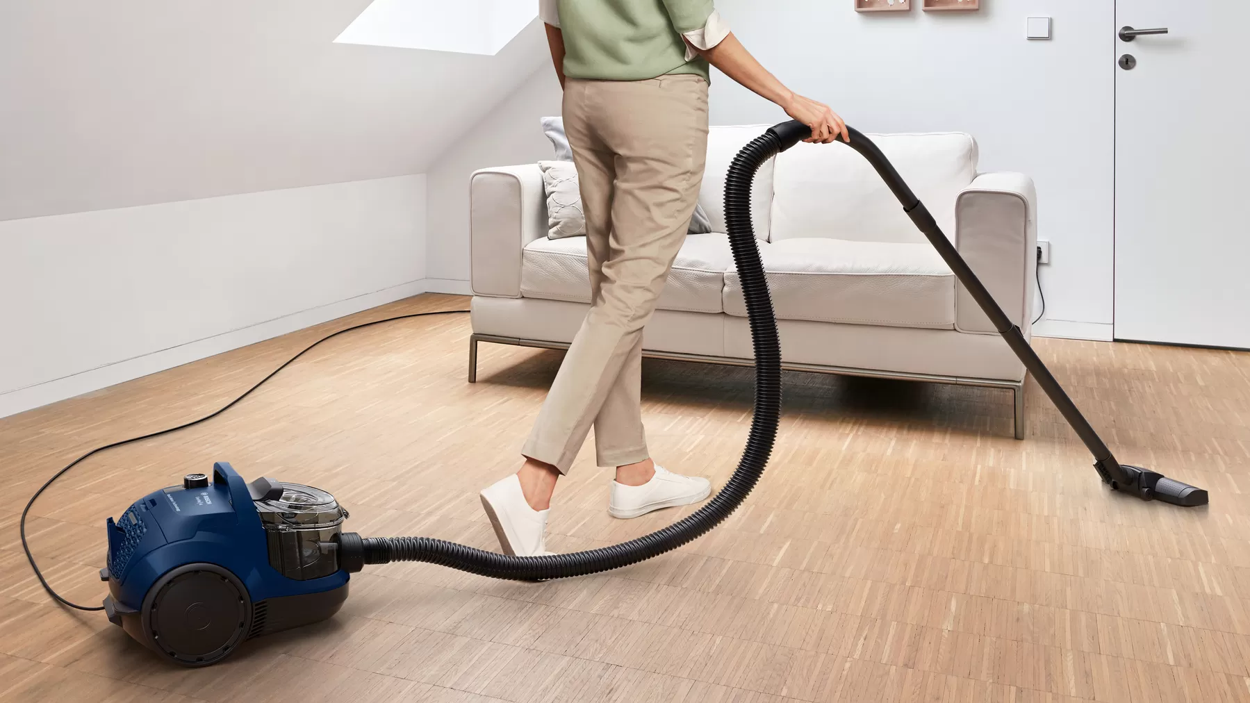 Woman vacuuming hardwood floor with blue vacuum cleaner, white shoes and beige pants.