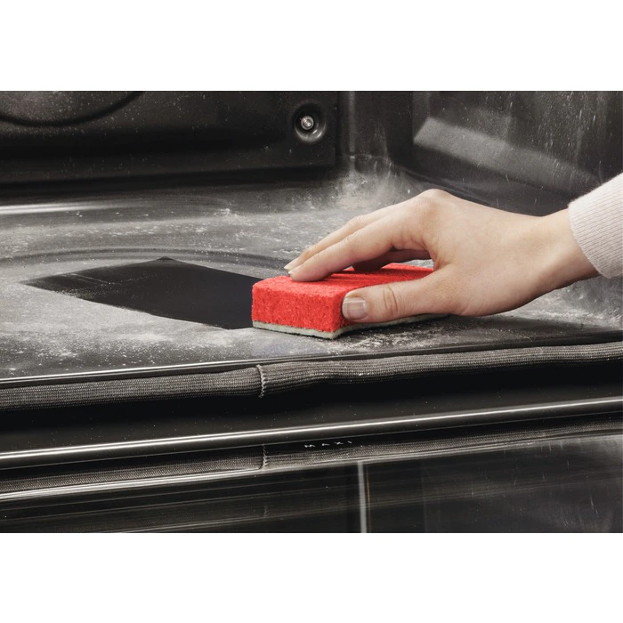 Een hand reinigt een vuile oven met een rode spons. Ovenrekken en glas zijn zichtbaar.