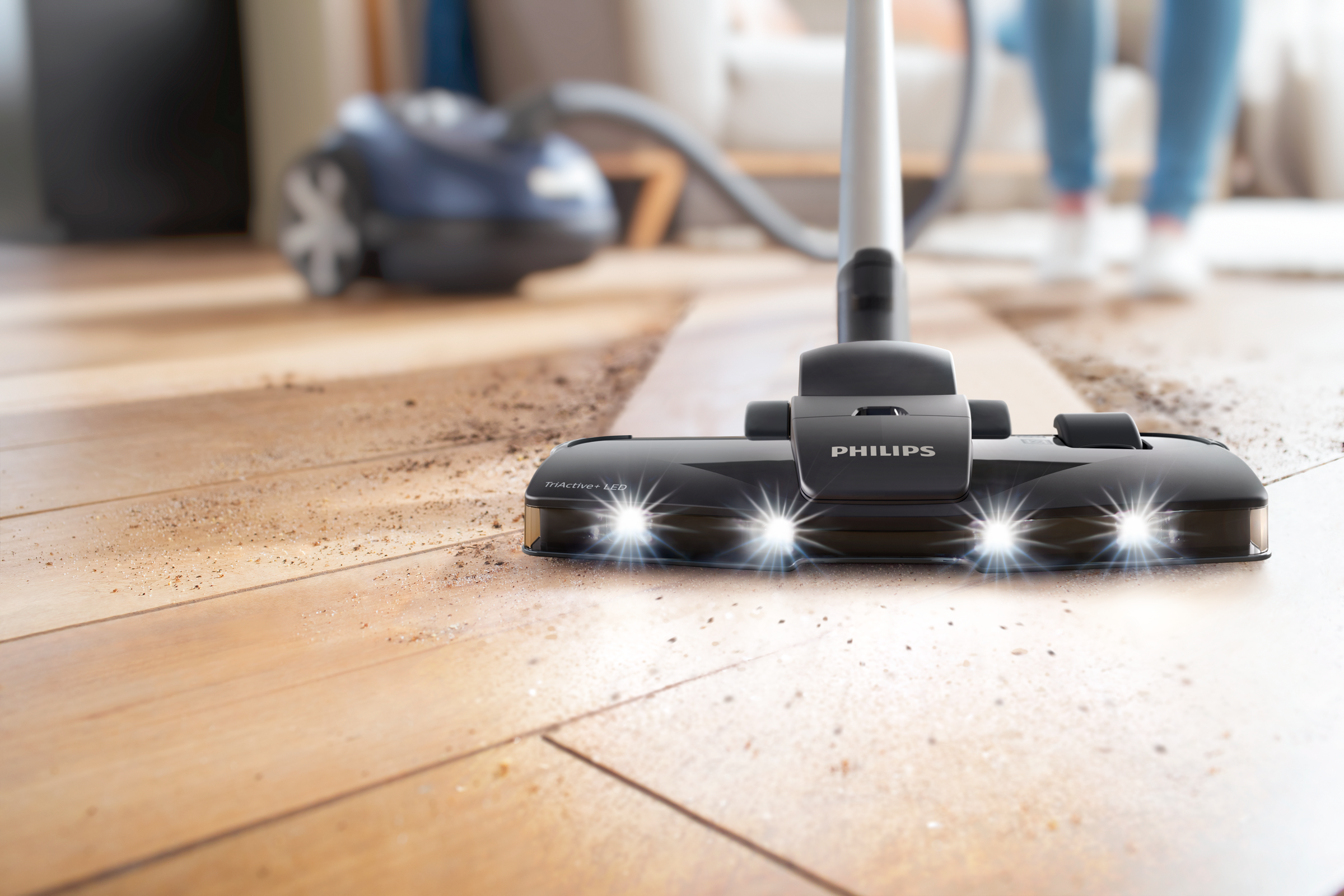 Een zwarte Philips stofzuigerkop op een houten vloer, met heldere lichten. Een persoon staat op de achtergrond.