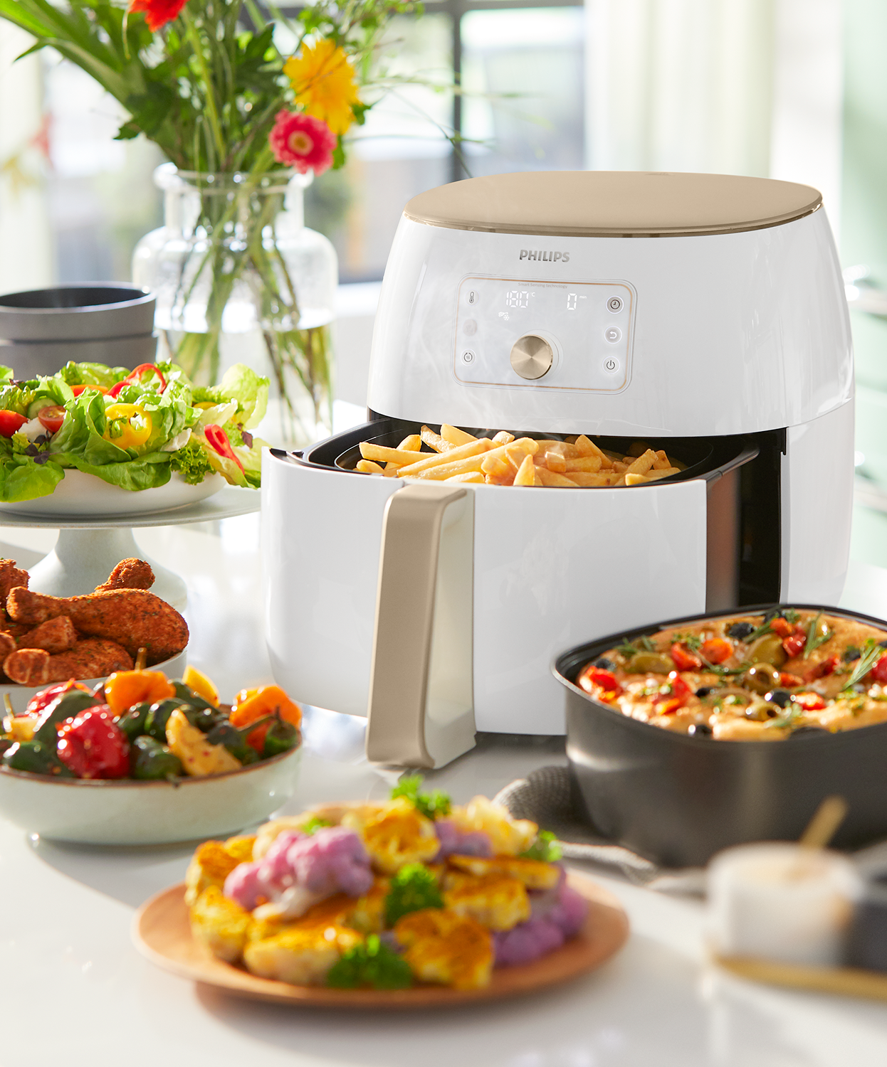 Air fryer with fries, other cooked food, flowers in background on a white table.