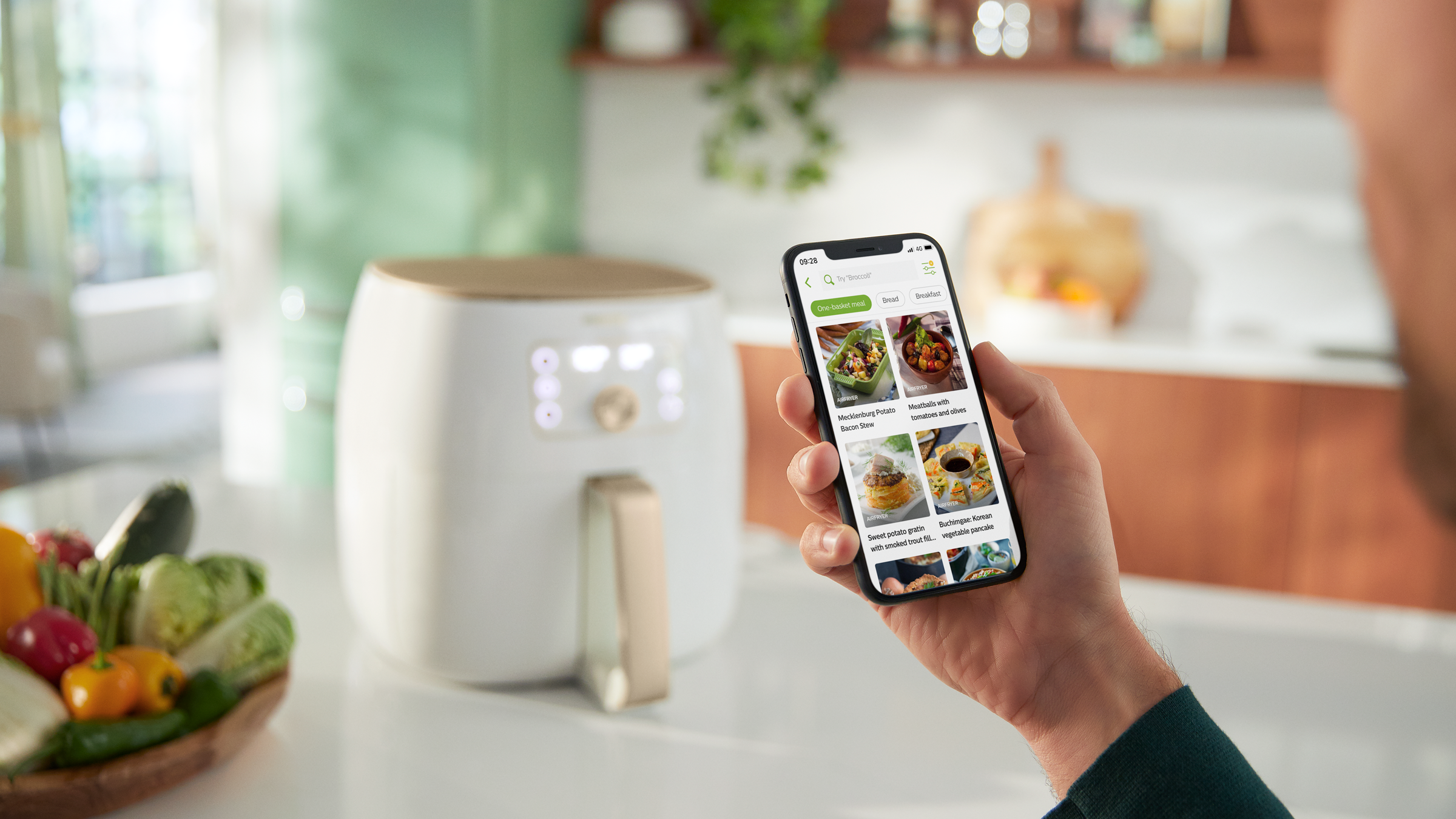Person holding phone with food app. Air fryer and ingredients on the background.