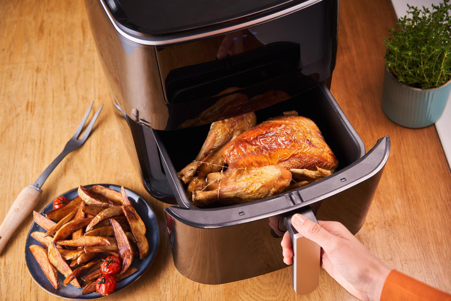 Heißluftfritteuse mit gebratenem Hähnchen und Pommes, Person hält Griff, Holztisch.