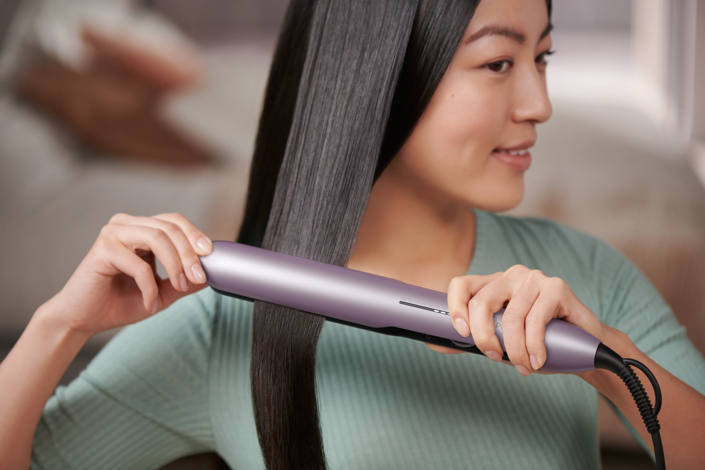 Woman with straight hair using a hair straightener. She is in a green top, and the background is blurred.