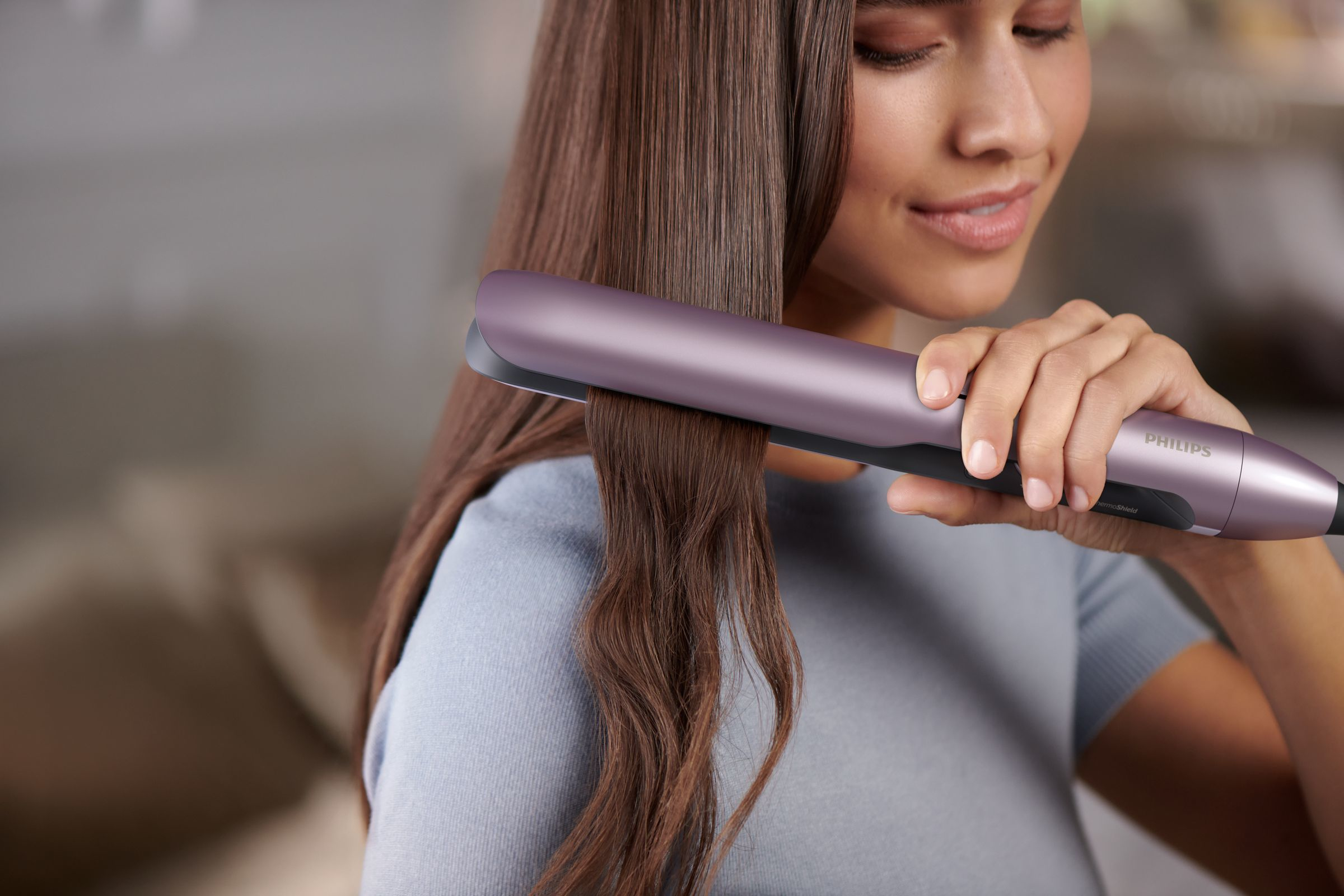 A woman using a purple hair straightener on her brown hair. She is smiling.