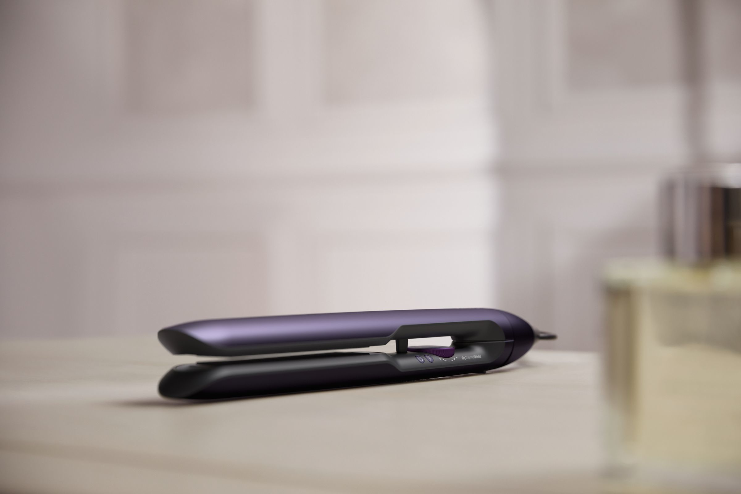 A purple and black hair straightener is on a wooden surface. A bottle is blurred in the background.