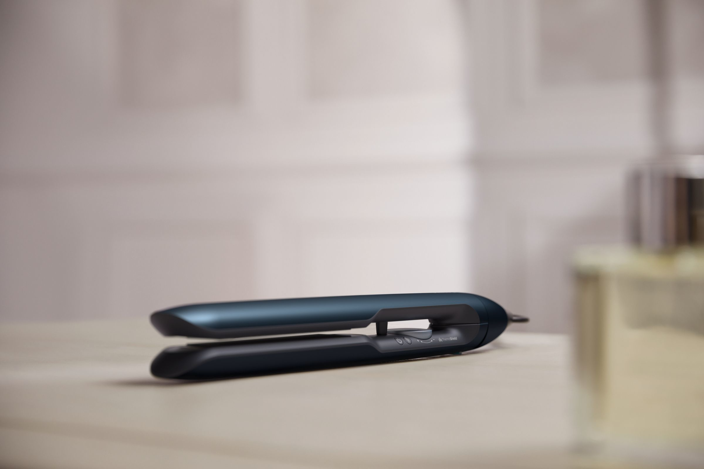 A dark blue flat iron lies open on a wooden surface, with a blurred perfume bottle.