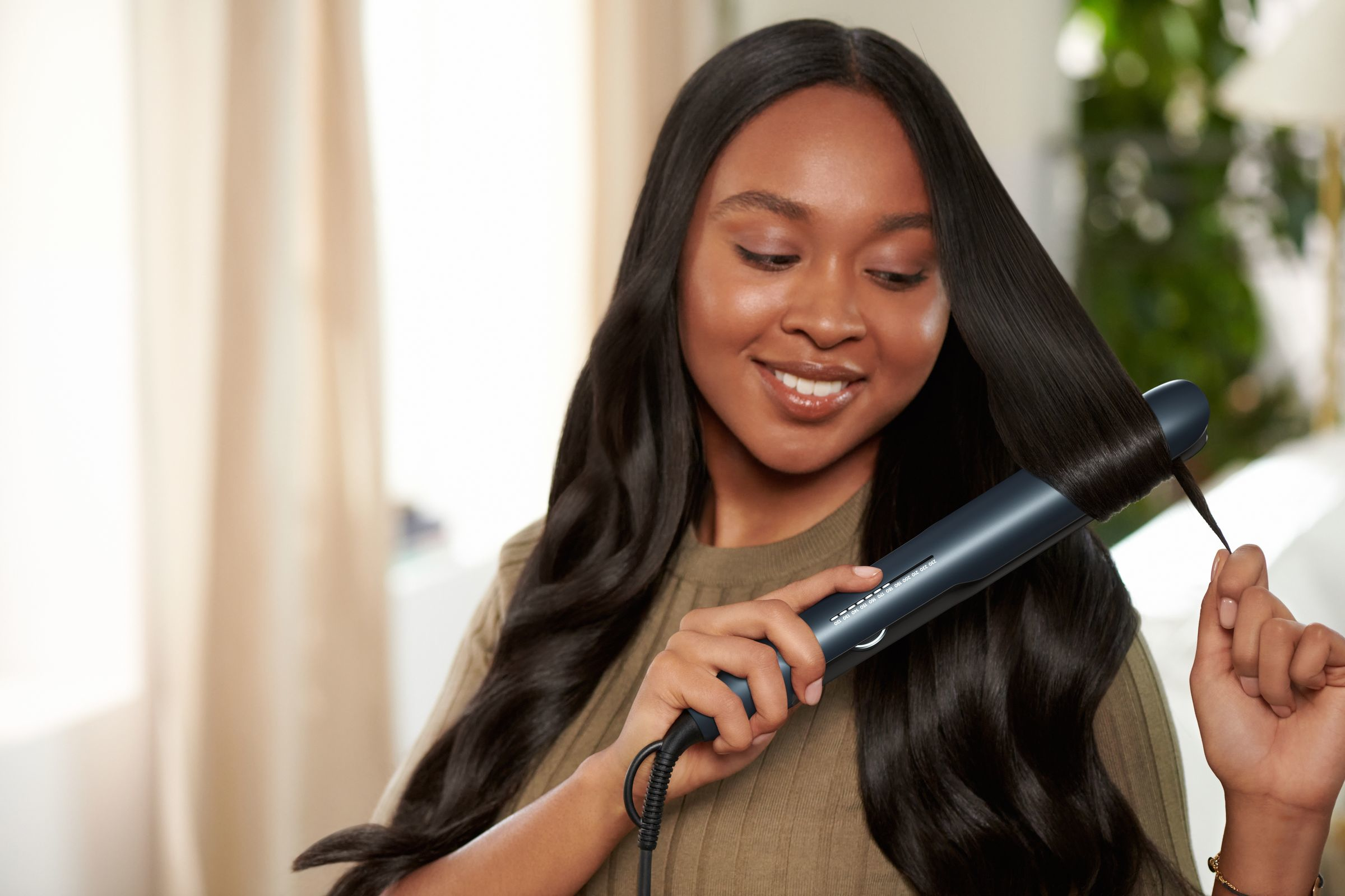 A woman styling her long dark hair with a hair straightener, smiling at the camera.