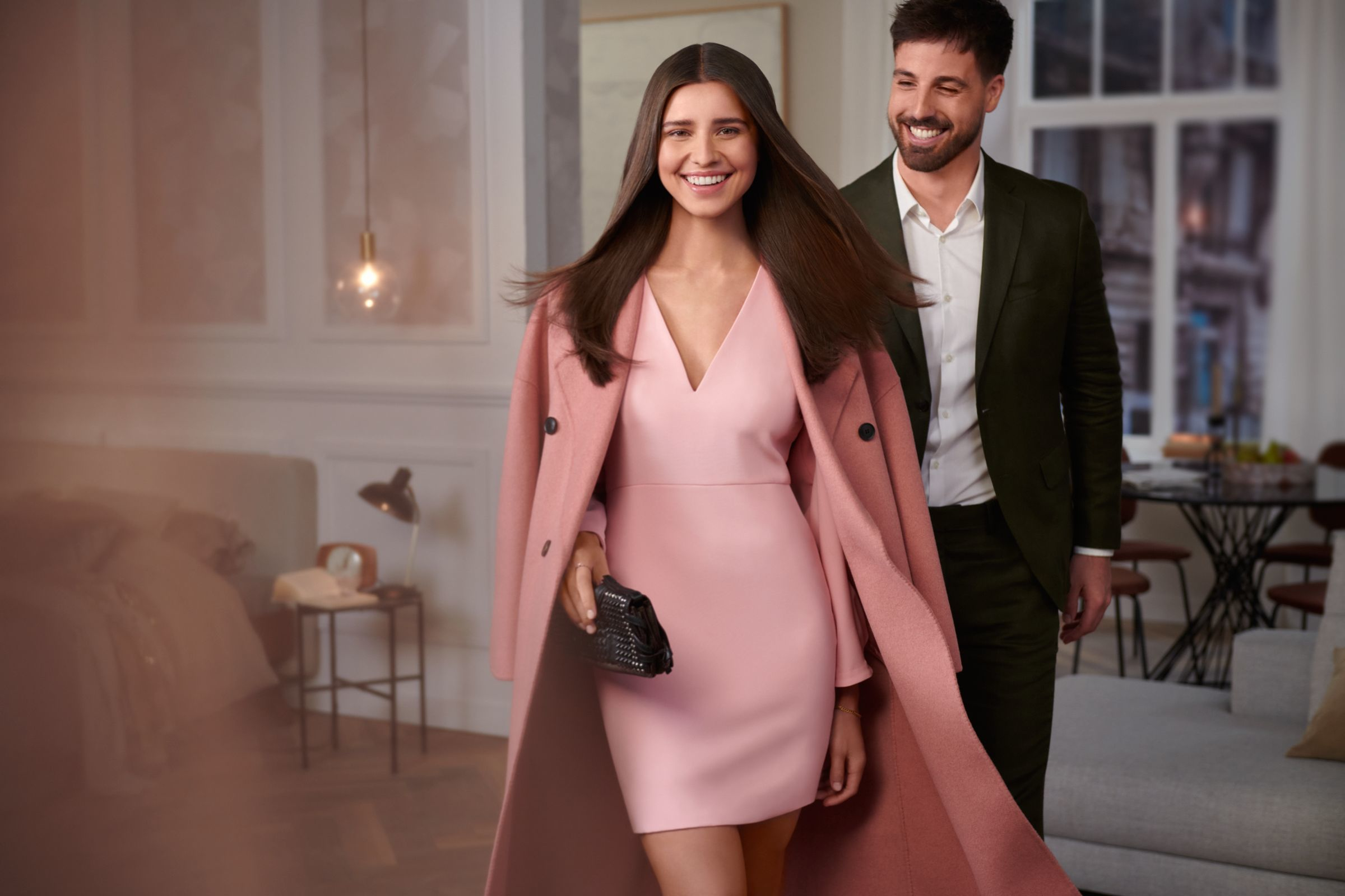 A woman in pink dress and coat walks with a man, with a modern interior background.