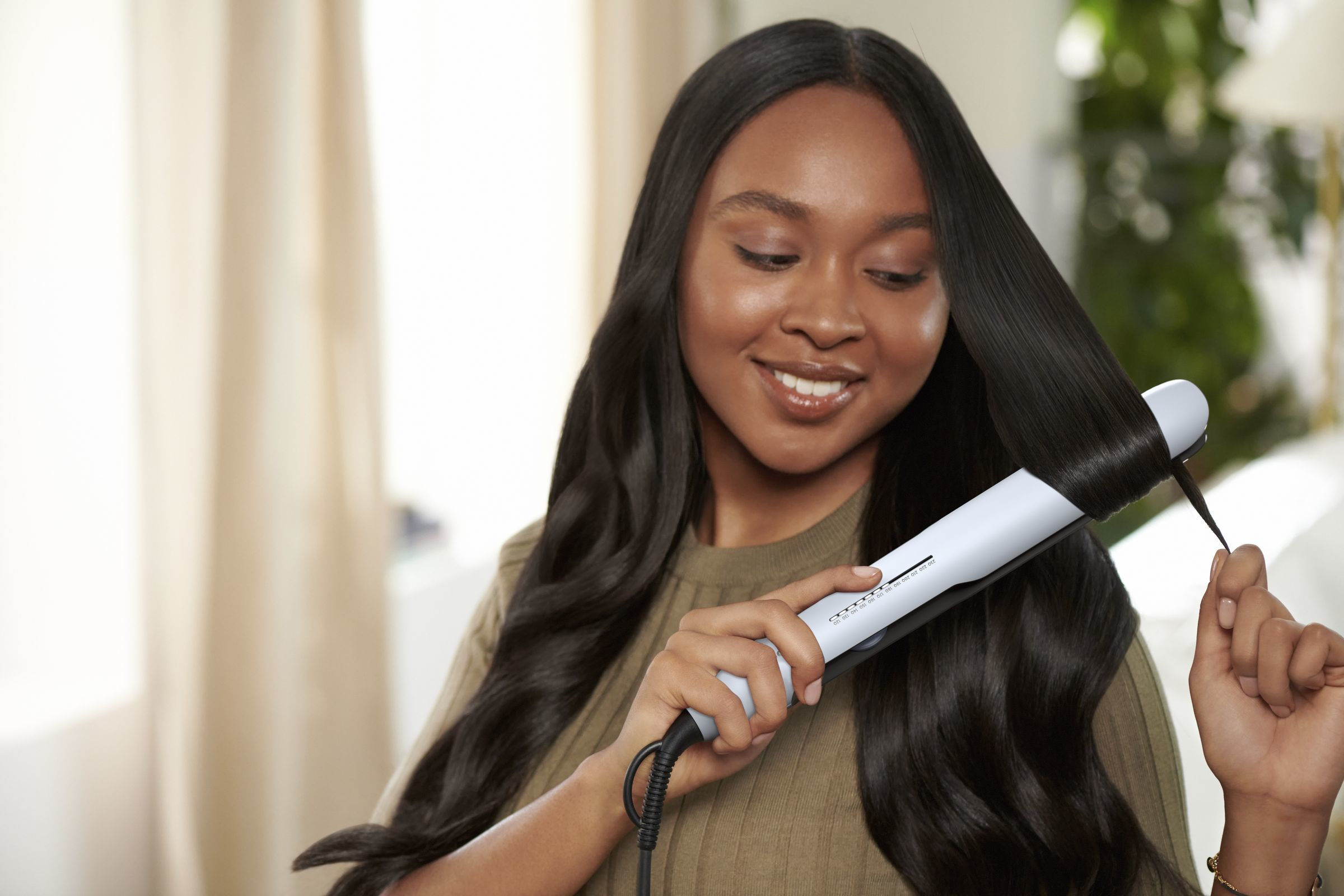 Woman with long dark hair using a hair straightener, smiling, indoors, with curtains and lamp.