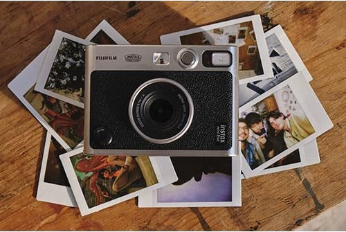 A silver camera with Instax on it is surrounded by photos on a wood table.