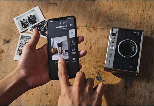 A person's hands holding a phone displaying a photo, with a camera and photos on a table.