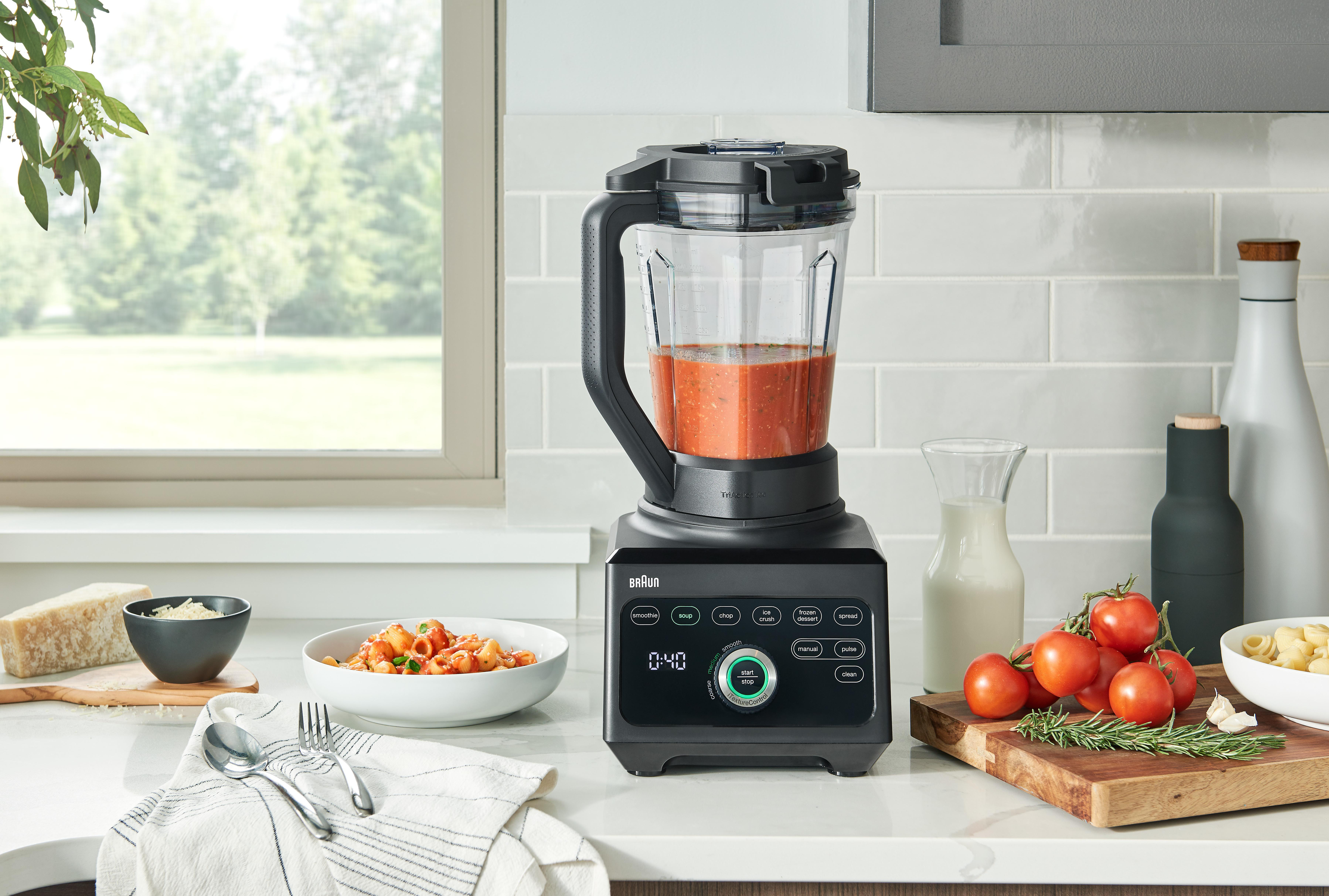 Schwarzer Mixer auf einer Theke, mit einer Schüssel Pasta und Tomaten. Ein Fenster befindet sich im Hintergrund.