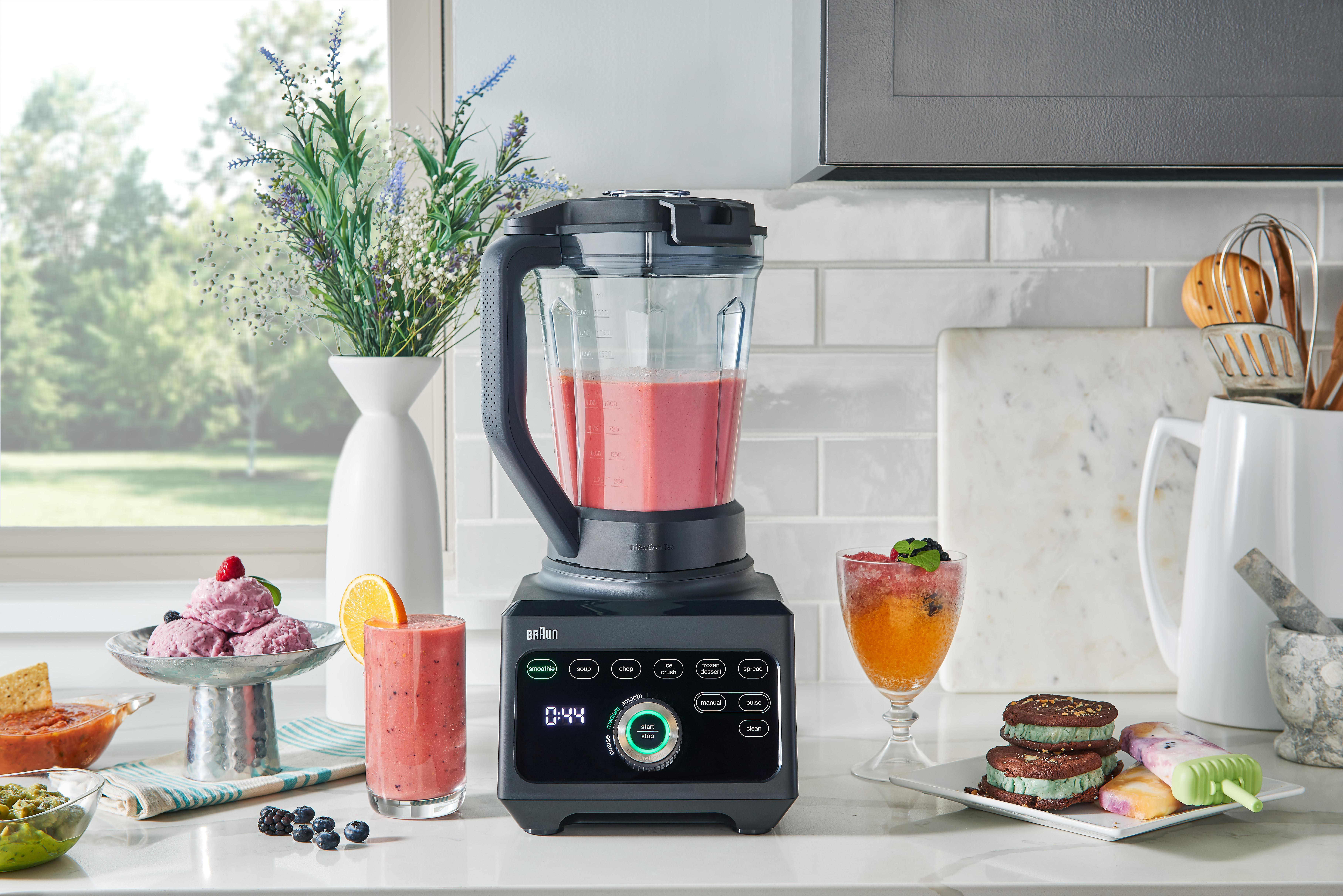Mixer mit rosa Smoothie, umgeben von anderen Getränken und Desserts auf weißer Theke.