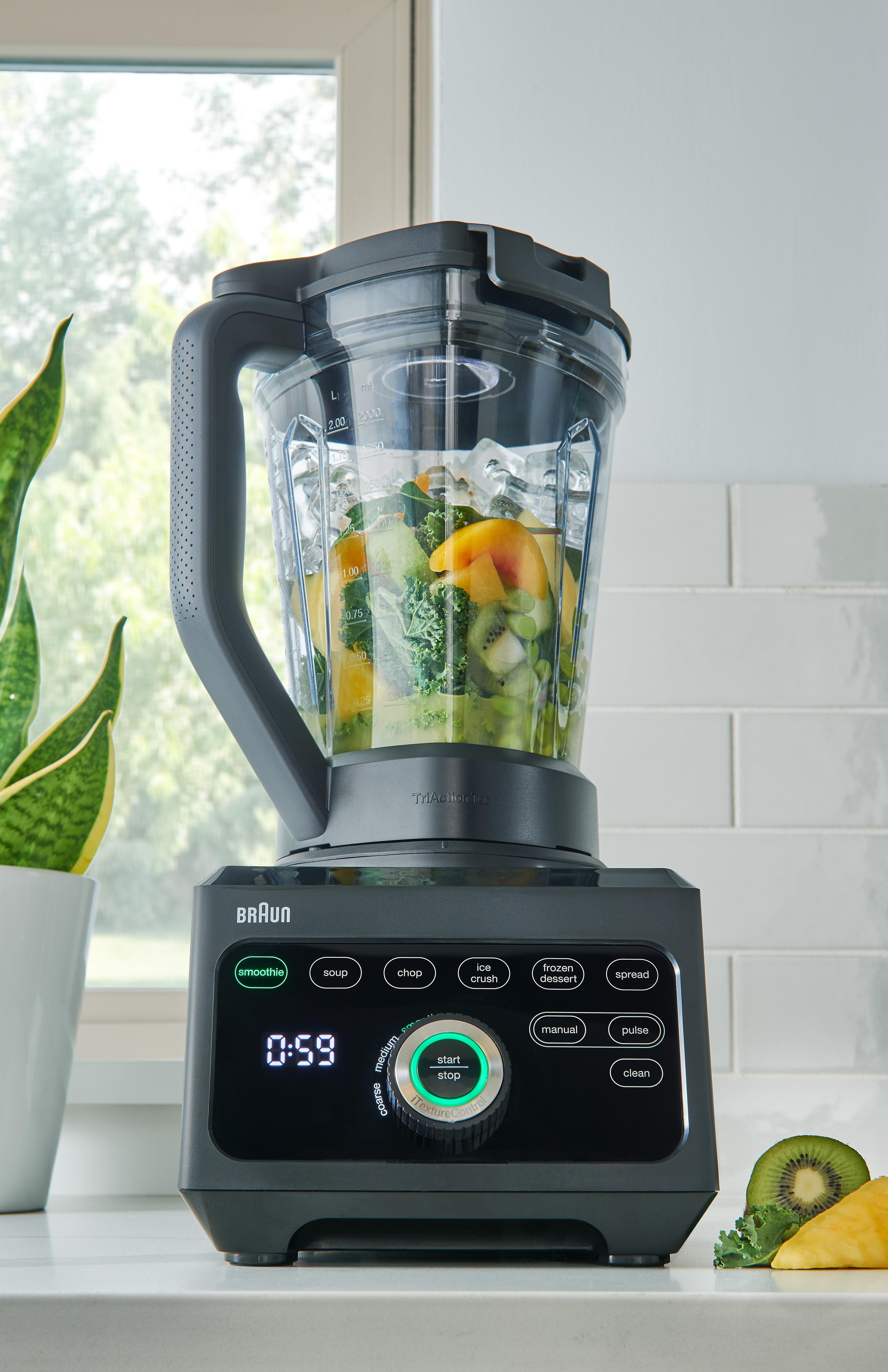 Mixer mit Obst und Gemüse, auf weißer Theke, nah an einem Fenster.