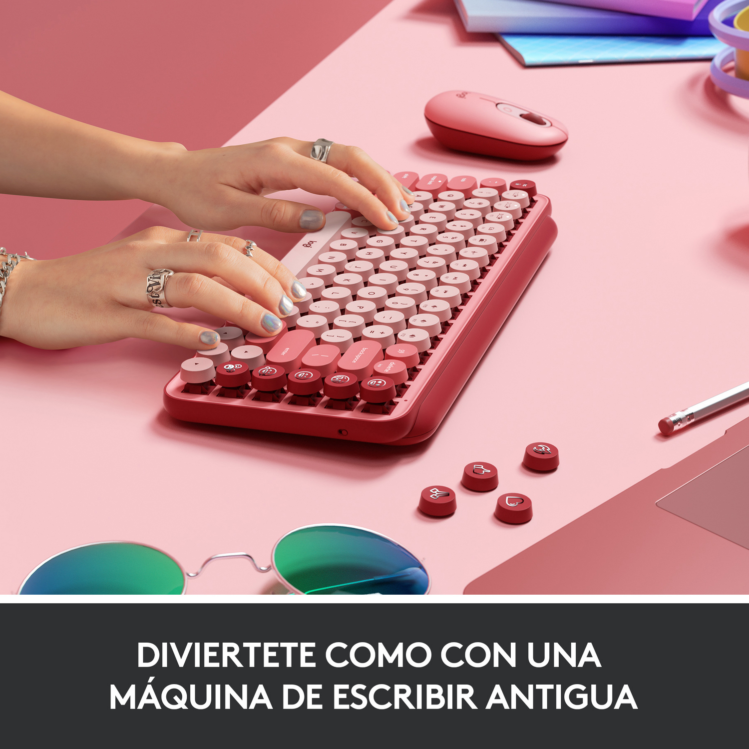 Manos escribiendo en teclado rosa, ratón a la derecha, gafas delante. Fondo rosa.