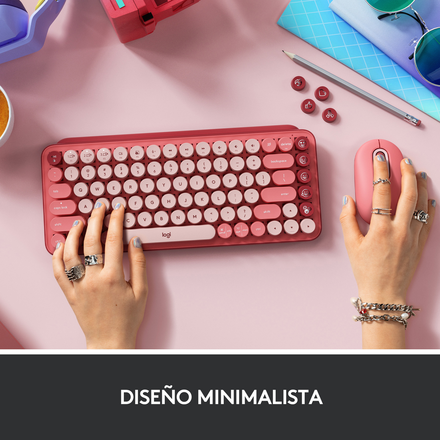 Manos en un escritorio rosa con teclado, ratón y accesorios rojos.