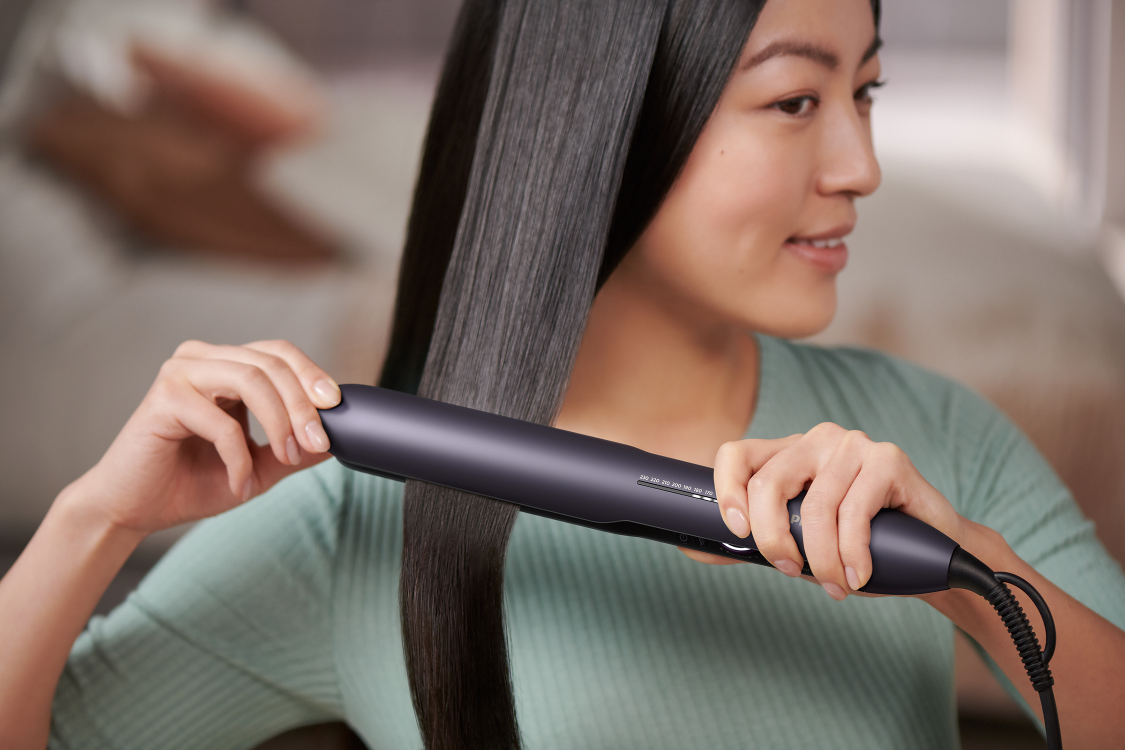 A woman straightens her hair with a flat iron. She is wearing a green shirt.
