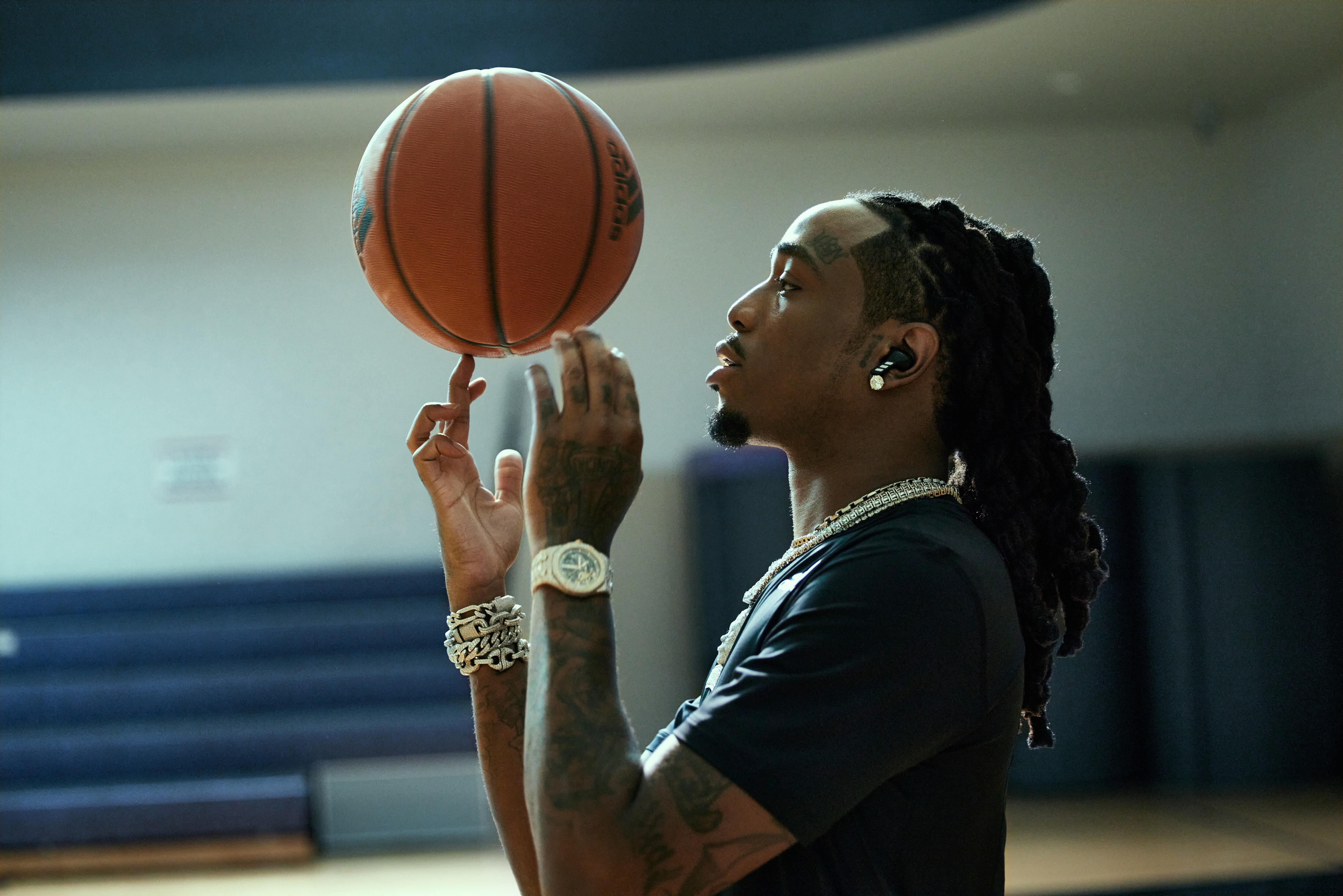 Ein Mann balanciert einen Basketball auf dem Finger, trägt Schmuck und Ohrhörer, drinnen.