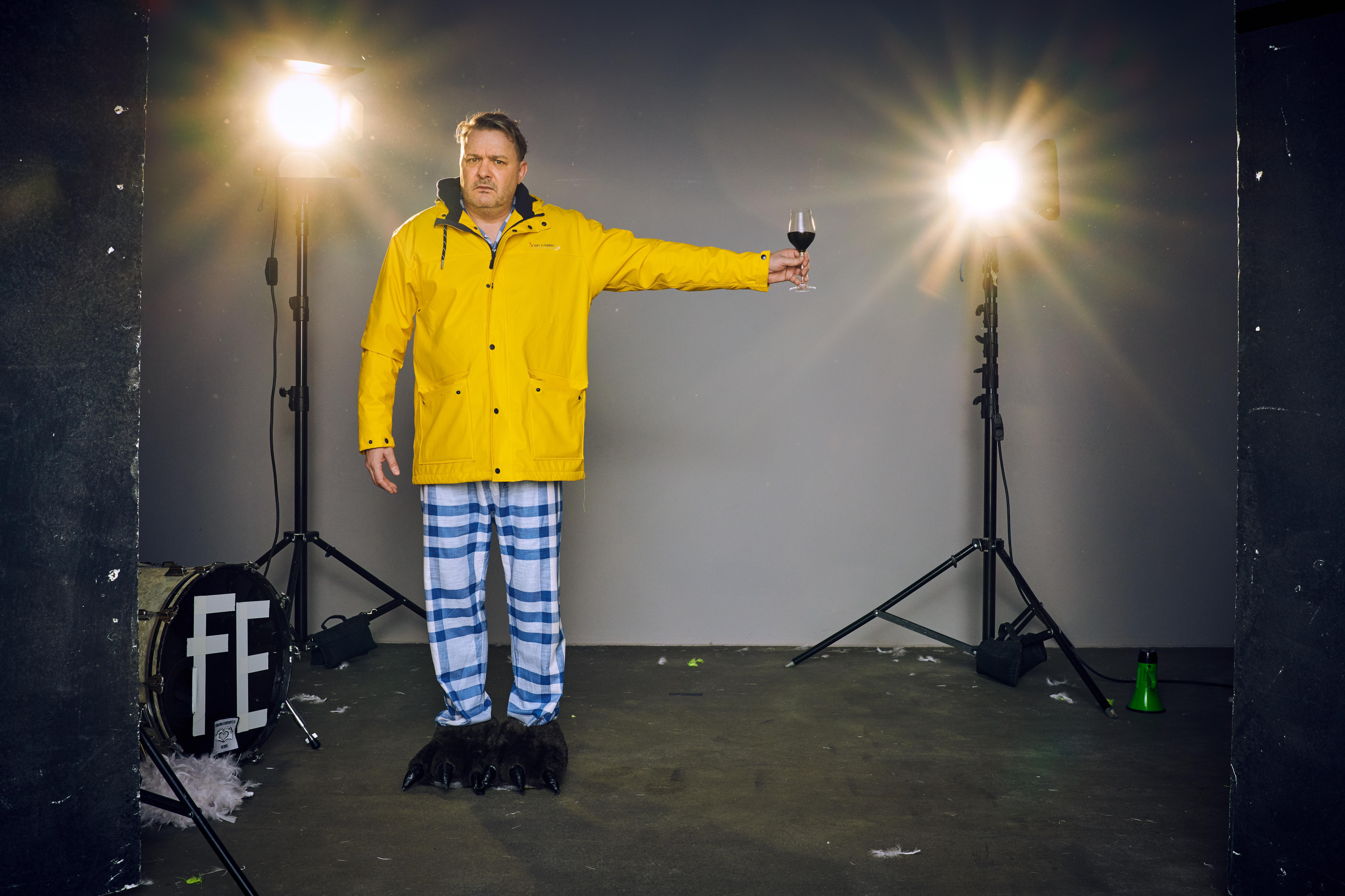 Mann in gelbem Mantel, Pyjamahose und Bärenfüßen, hält Weinglas, Studiolichter im Hintergrund.