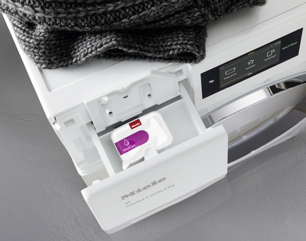 Machine à laver avec tiroir à détergent ouvert contenant une dosette WoolCare et un tricot gris.
