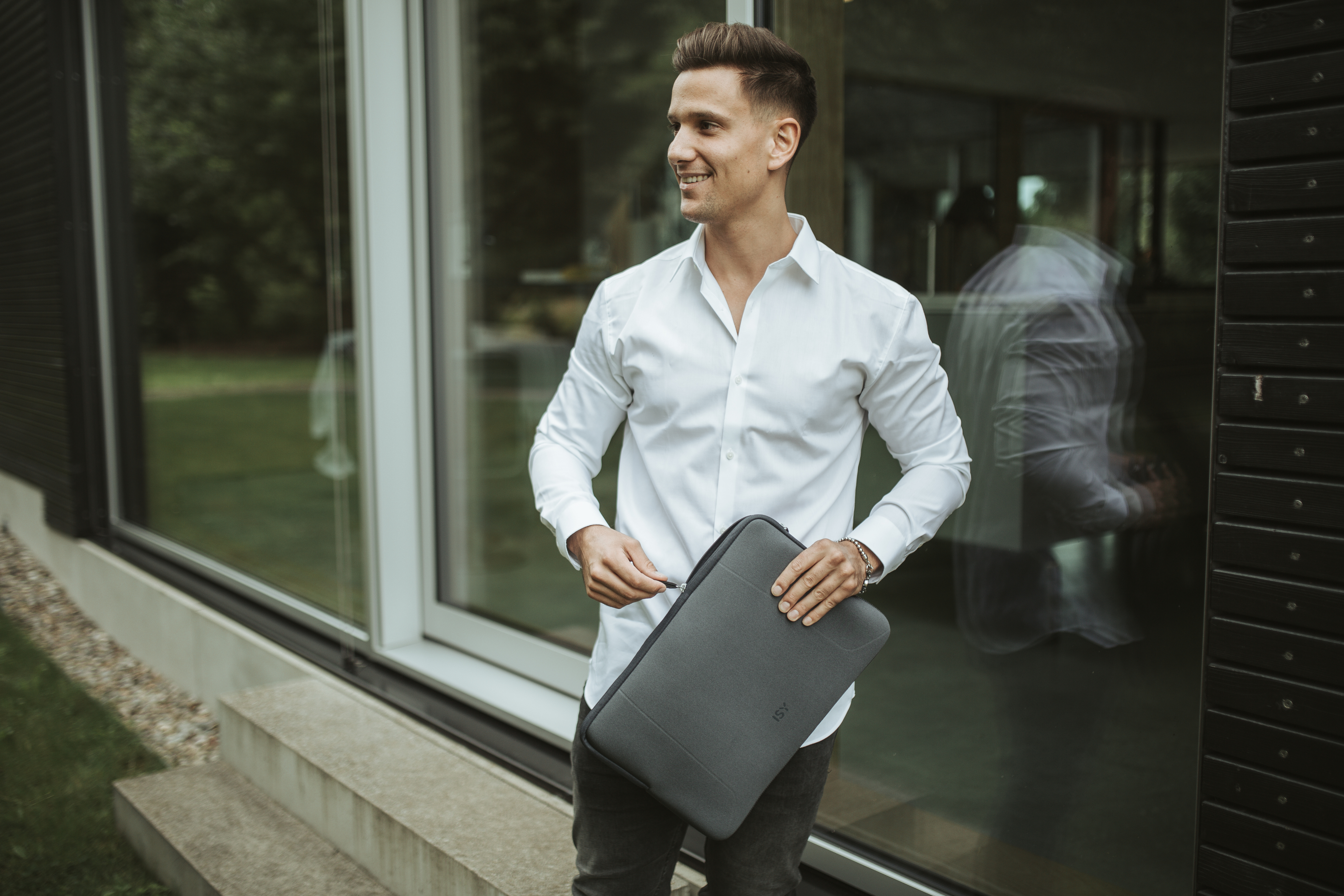 Homme en chemise blanche tenant une housse d'ordinateur portable grise, près d'une porte vitrée.