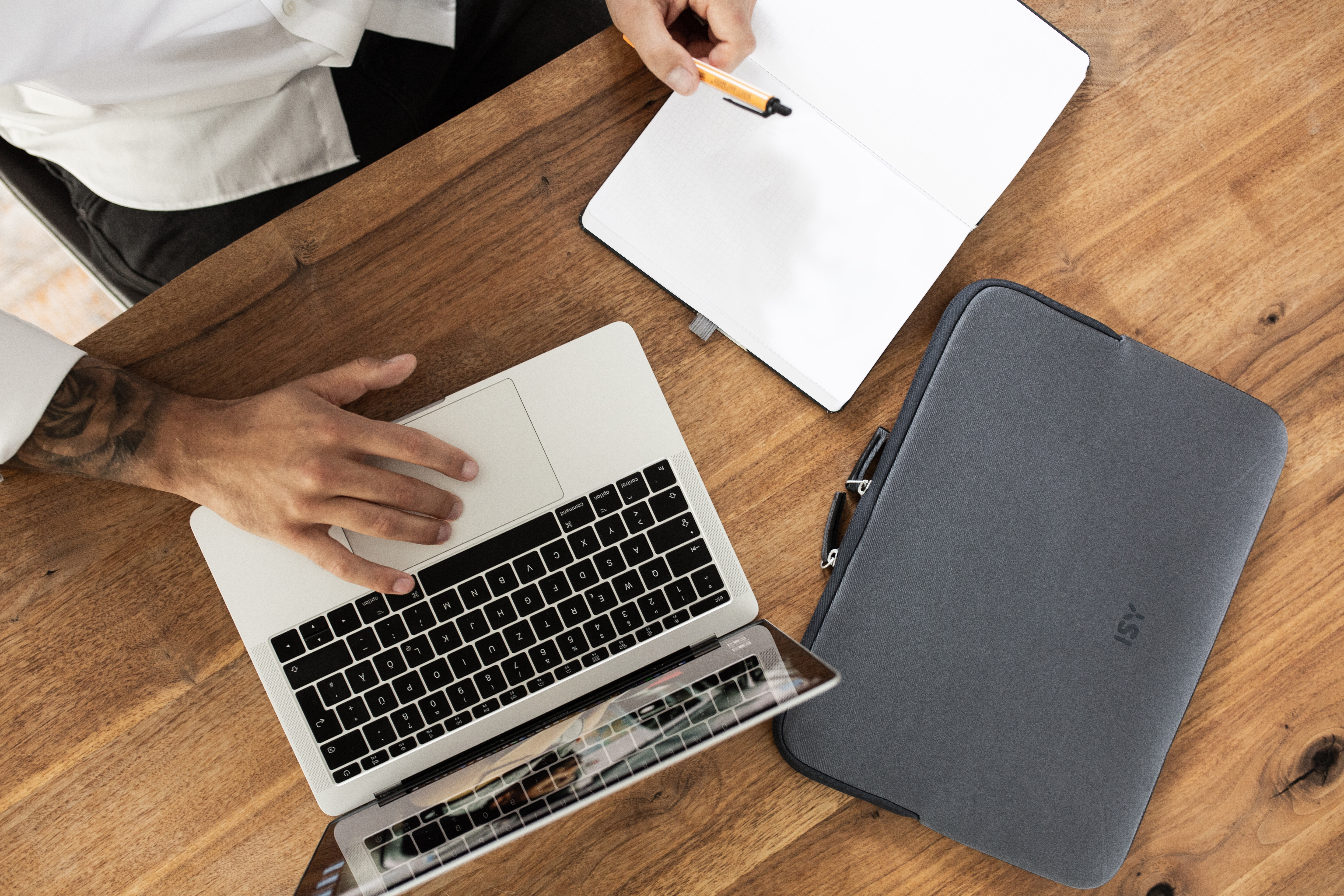 Les mains d'une personne travaillant sur une table en bois avec un ordinateur portable et un cahier. Une coque de téléphone est à proximité.