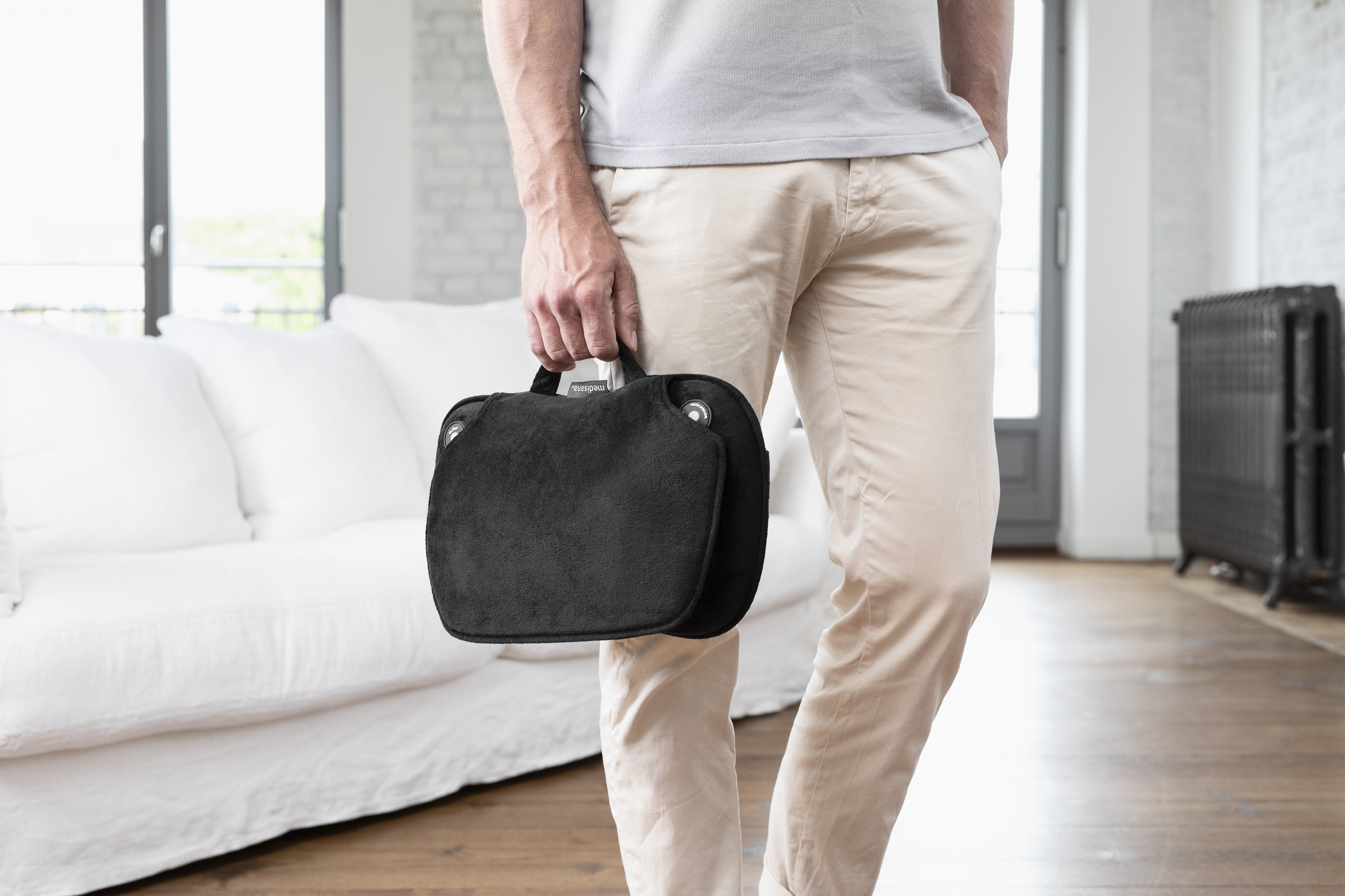 Un homme en pantalon beige tient un sac noir devant un canapé blanc. Plancher en bois.