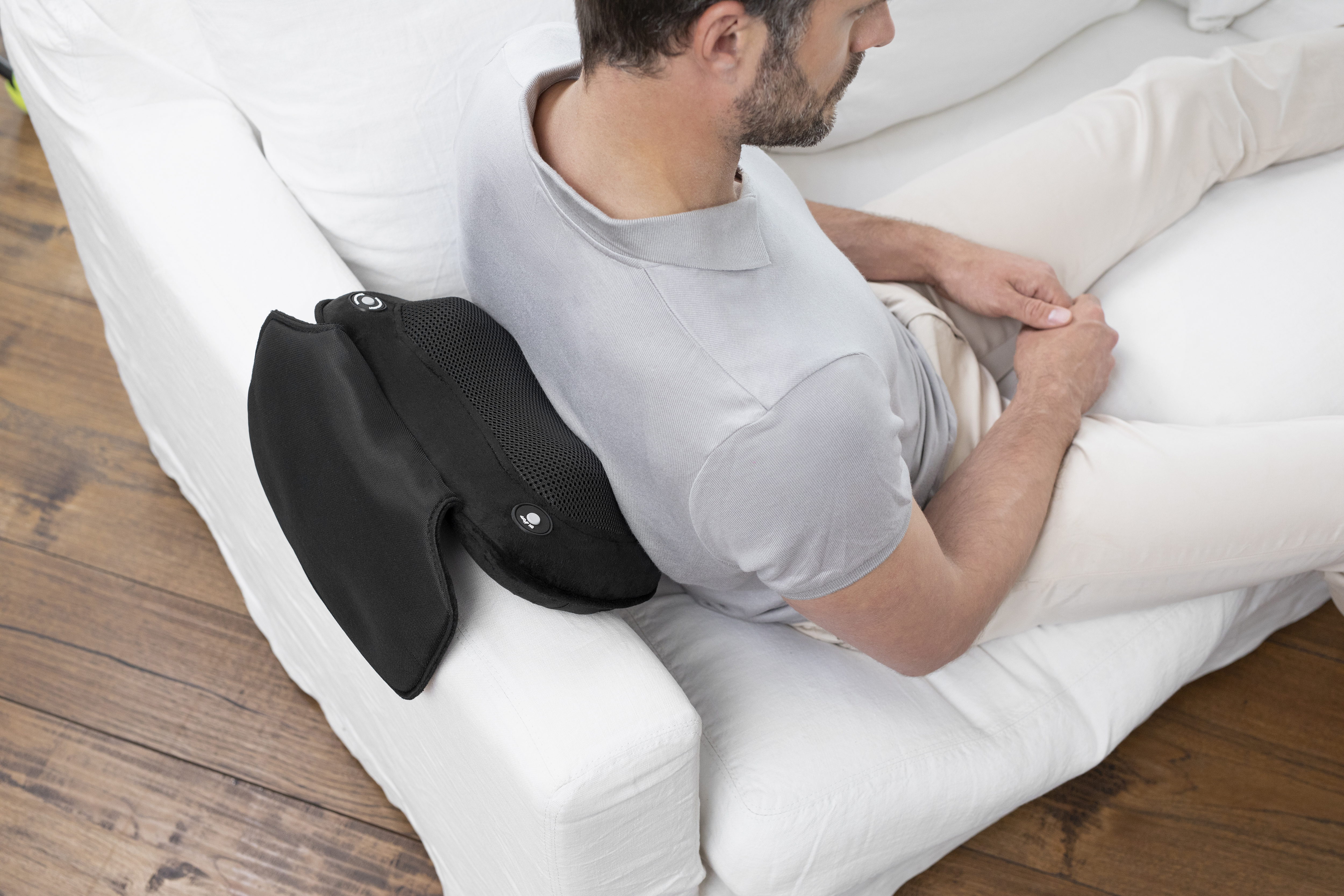 Un homme est assis sur un canapé blanc avec un coussin de soutien lombaire noir. Il porte une chemise grise et un pantalon beige.